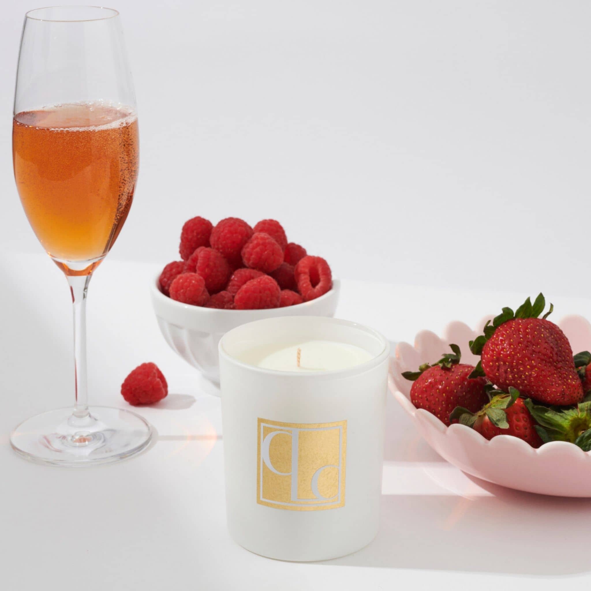 Glass of pink champagne, bowl of raspberries, and bowl of strawberries with a Caitlin Louise Collection candle on a white background