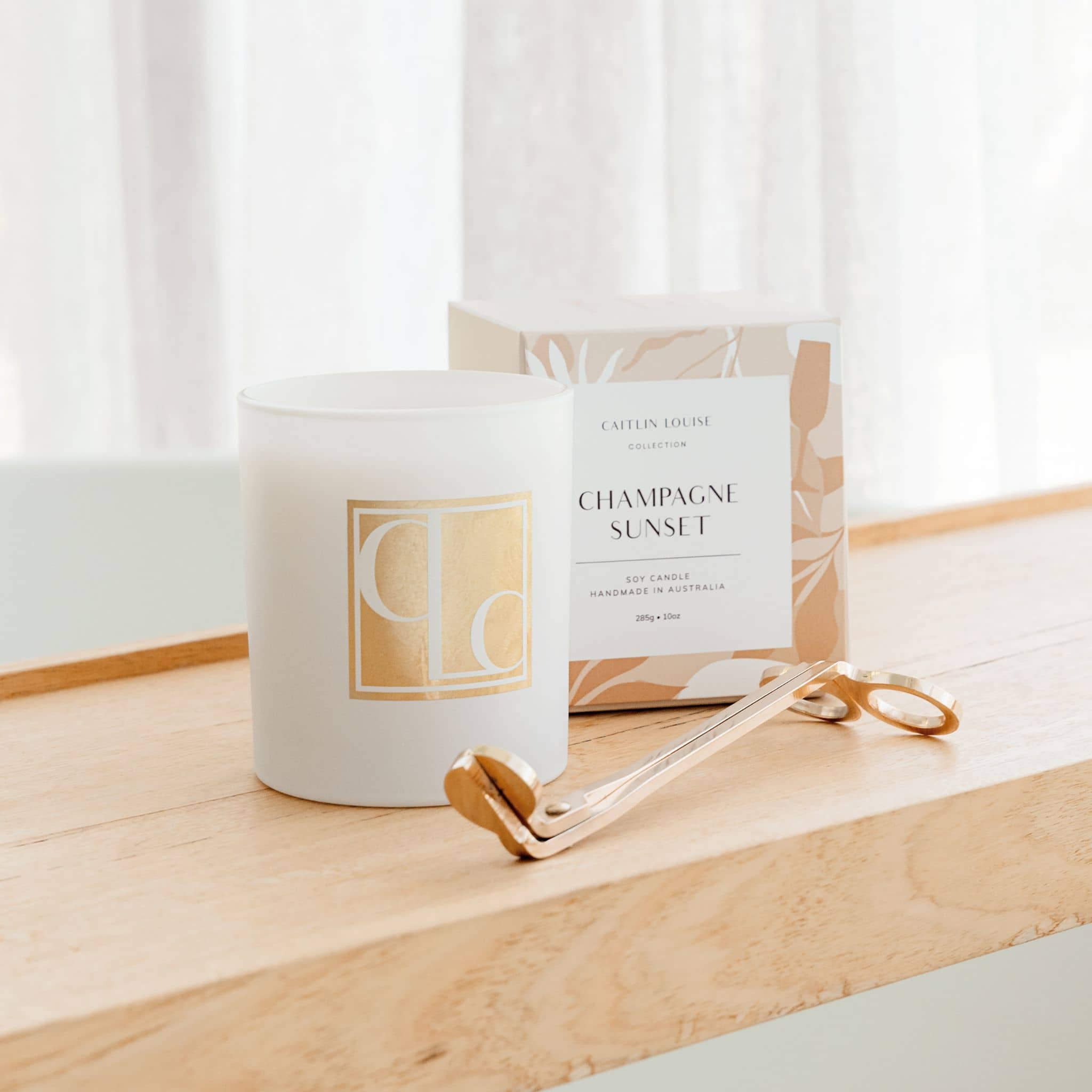 Candle labeled 'Champagne Sunset' with a decorative box and a gold wick trimer on a wooden surface.
