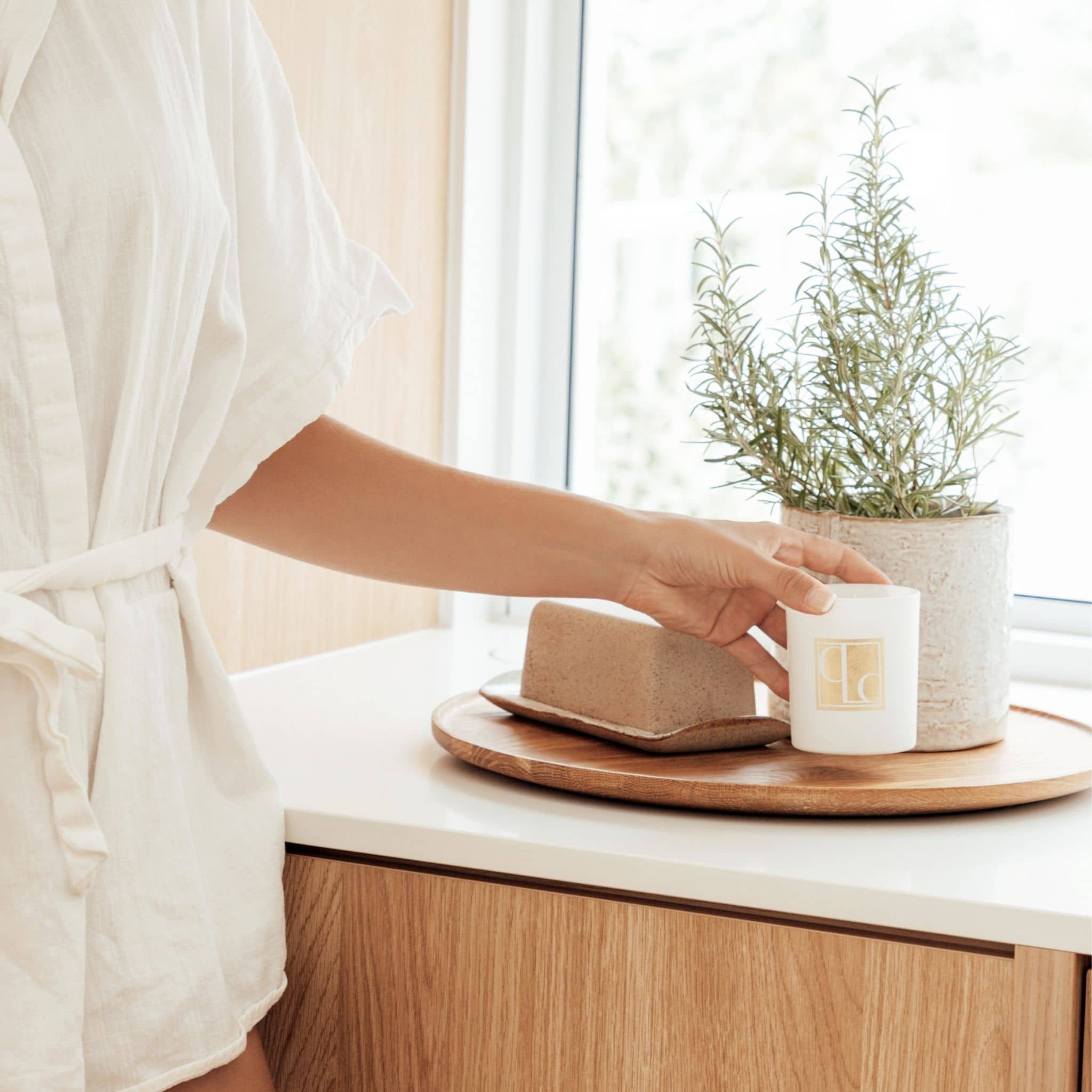 Person in a white robe holding a Caitlin Louise Collection candle with a plant and bread on a wooden tray.