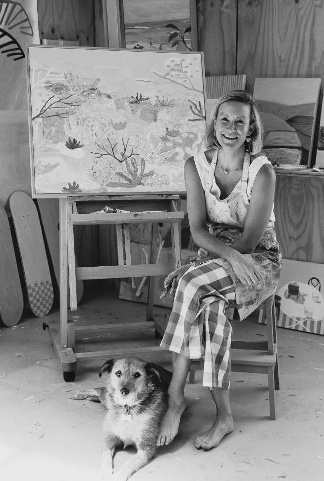 Woman sitting with a dog in an art studio surrounded by paintings and art supplies.