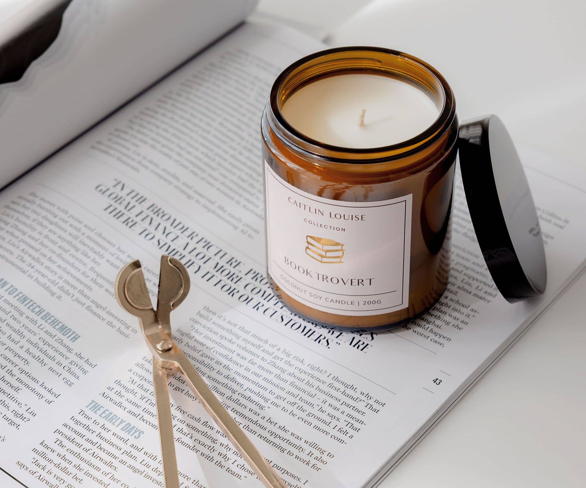 Candle in a glass jar labeled 'Booktrovert' on an open book with a pair of wick trimmers.