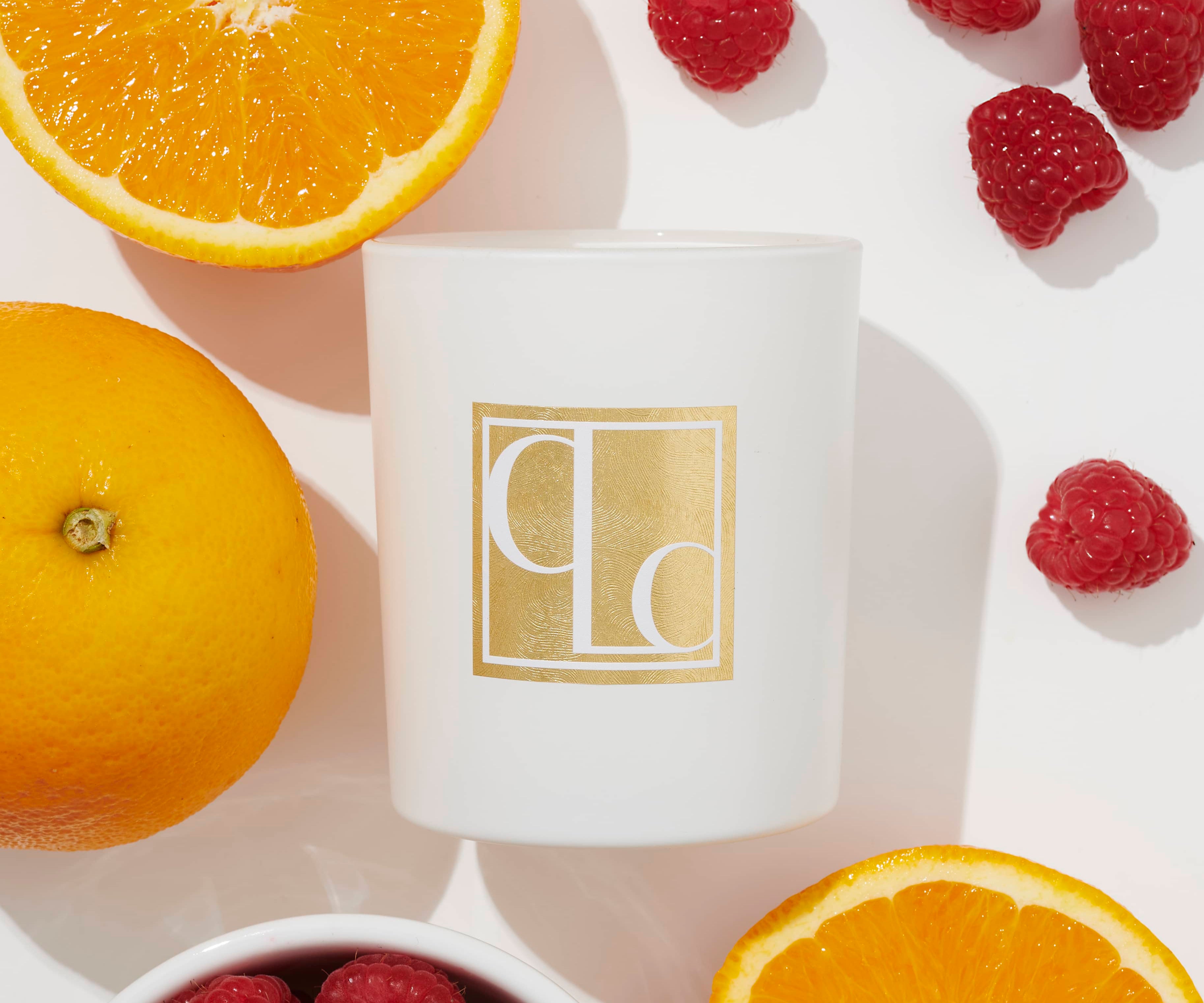 A white Caitlin Louise Collection candle with a gold label, surrounded by orange slices and raspberries, on a white background.