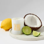 A white candle with the scent of coconut lime, placed on a marble surface, accompanied by a halved coconut, a lemon, and lime slices.