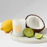 A white candle with the scent of coconut lime, placed on a marble surface, accompanied by a halved coconut, a lemon, and lime slices.