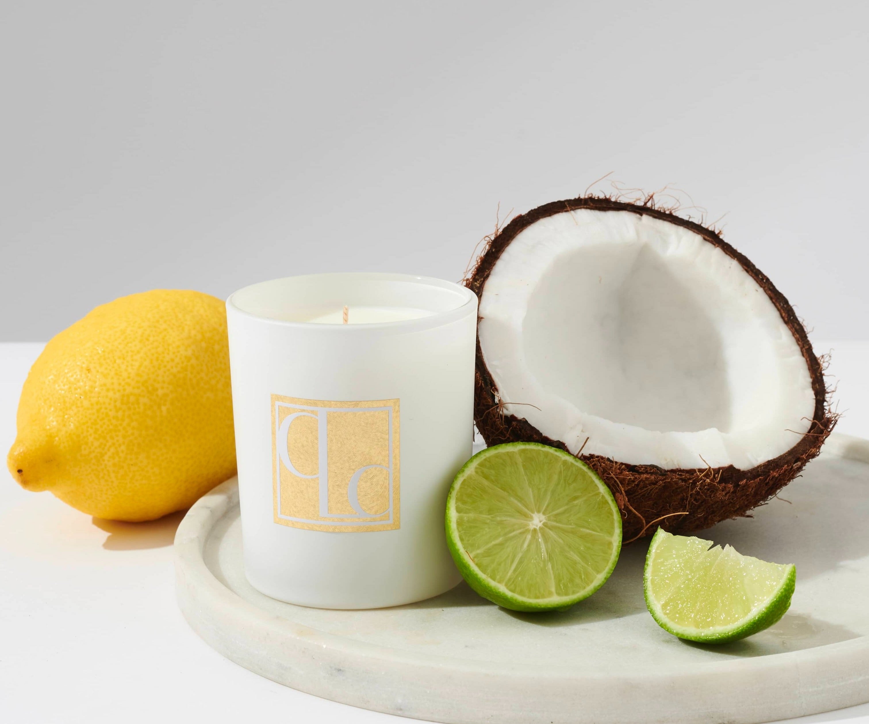 A white candle with the scent of coconut lime, placed on a marble surface, accompanied by a halved coconut, a lemon, and lime slices.