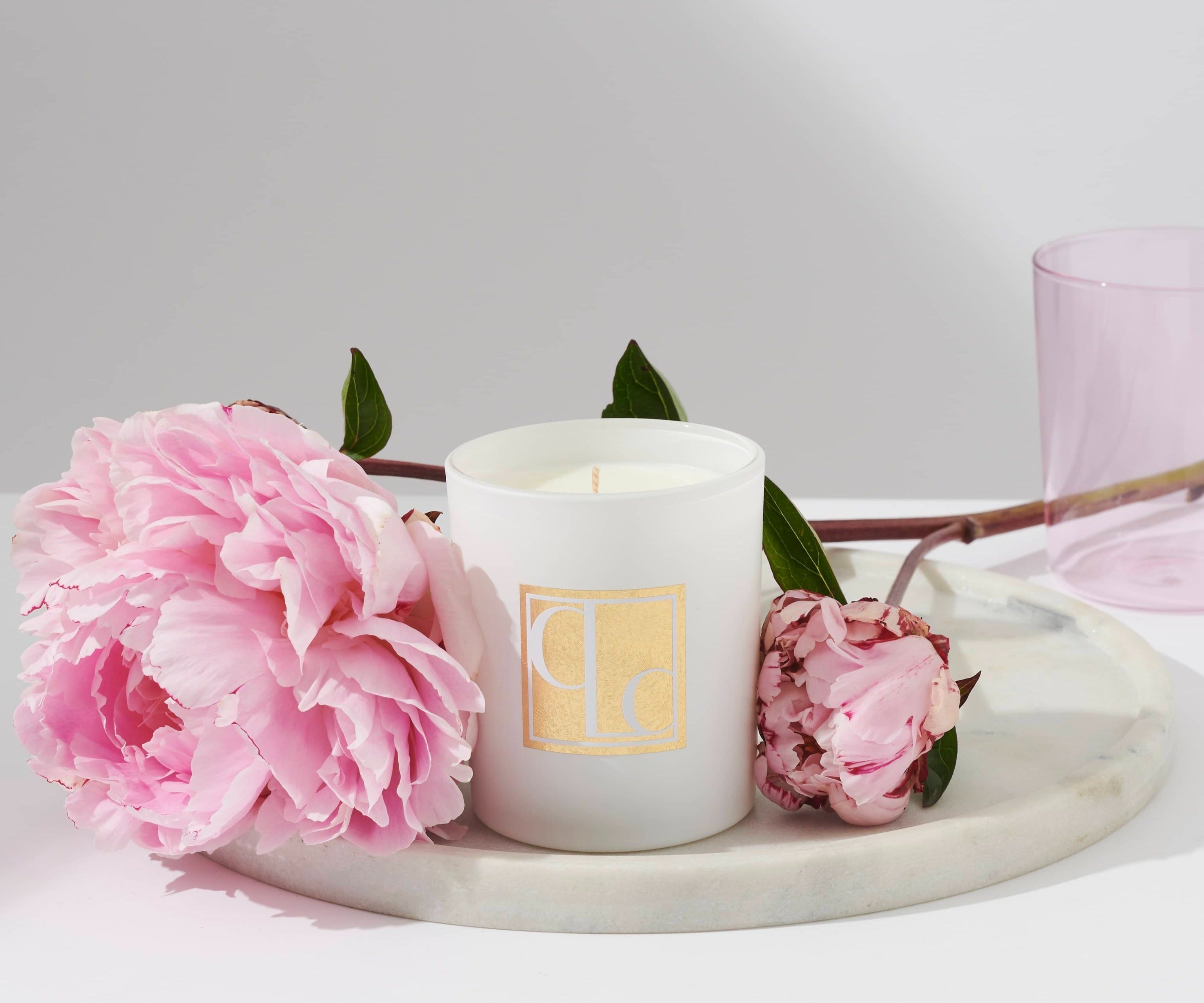 A white candle with the Lychee Peony scent, placed on a marble surface, surrounded by pink peony flowers and a pink glass.