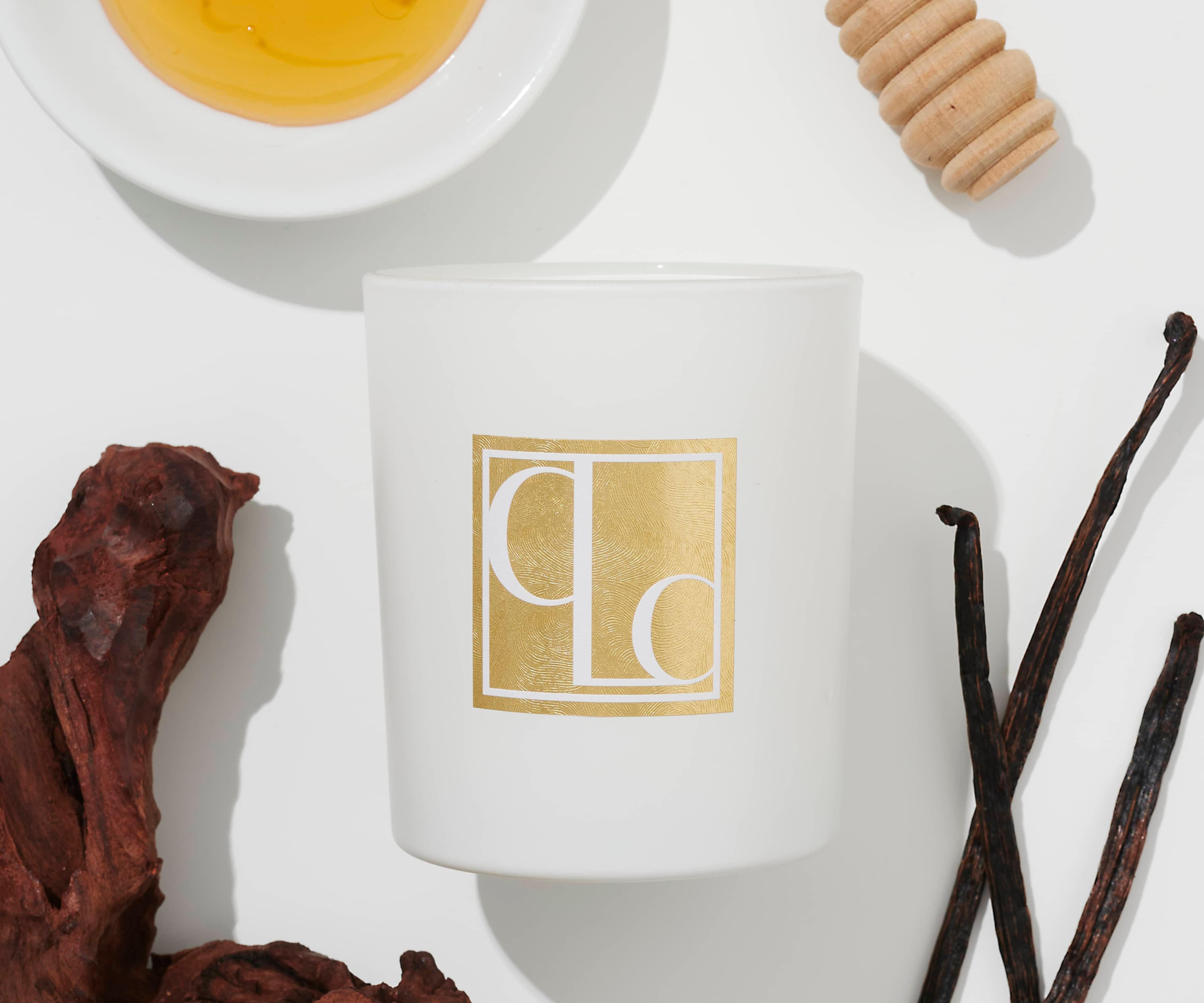 A white candle with the brand mark 'CLC' in gold, surrounded by honey, a honey dipper, dried fruits, and vanilla pods on a light background.