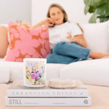 Caitlin Louise Collection's Patchouli and Sandalwood candle sitting on stack of books with woman on the lounge in the background.