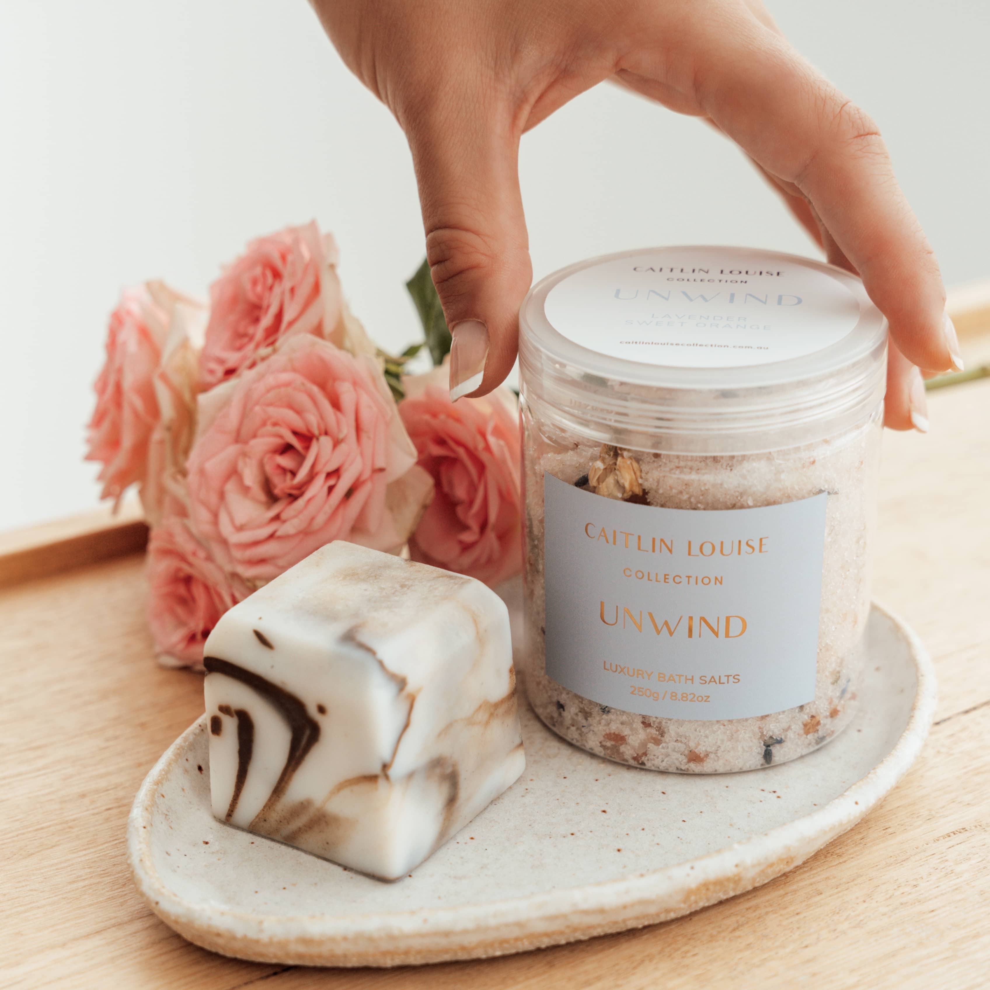 Hand reaching for Caitlin Louise Collection Unwind Luxury Bath salts, sitting on a tray with flowers behind.