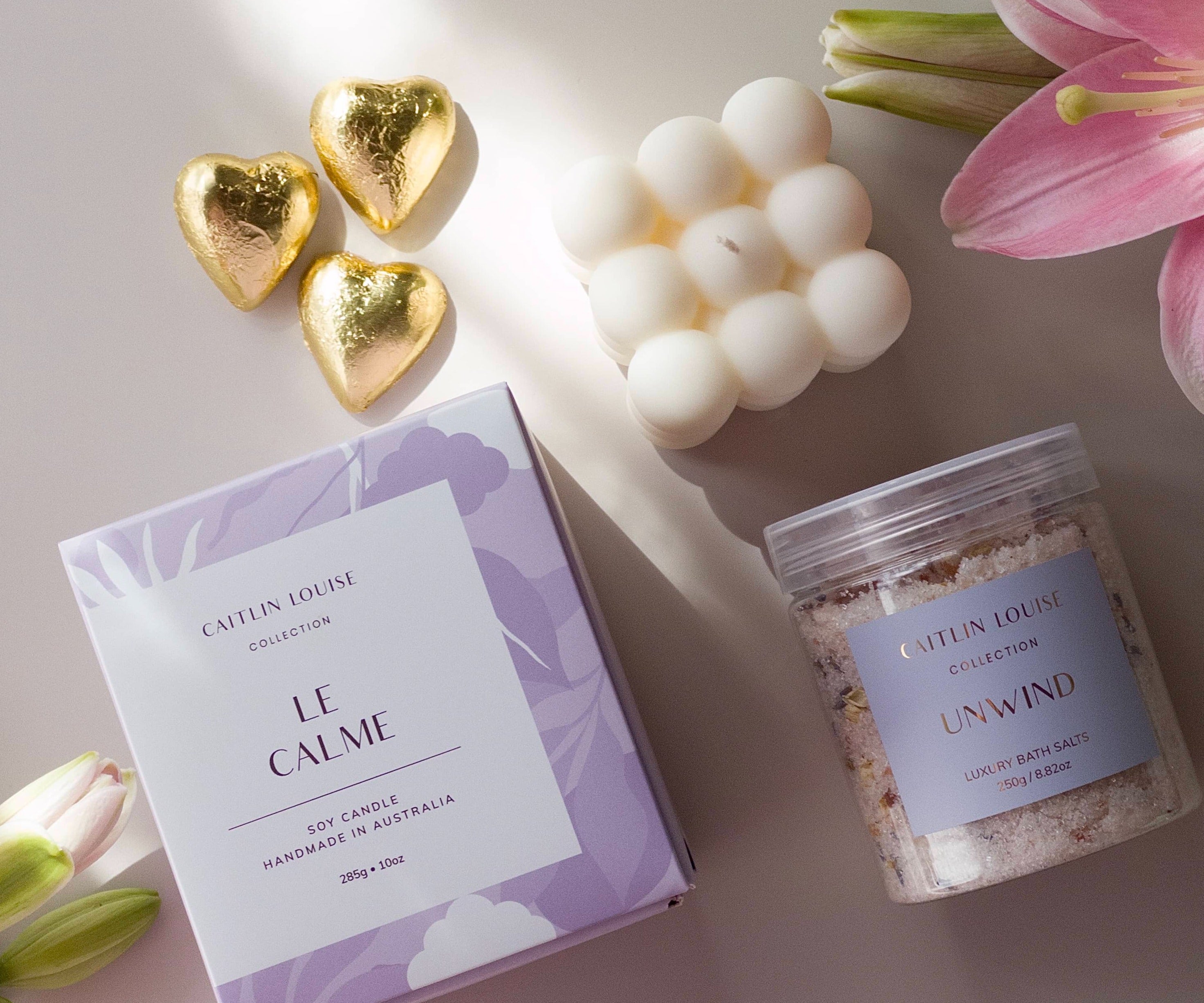 Flat lay of products in gift hamper - le calme scented candle, unwine bath salts, amara bubble cube candle and three heart chocolates - surrounded by flowers.