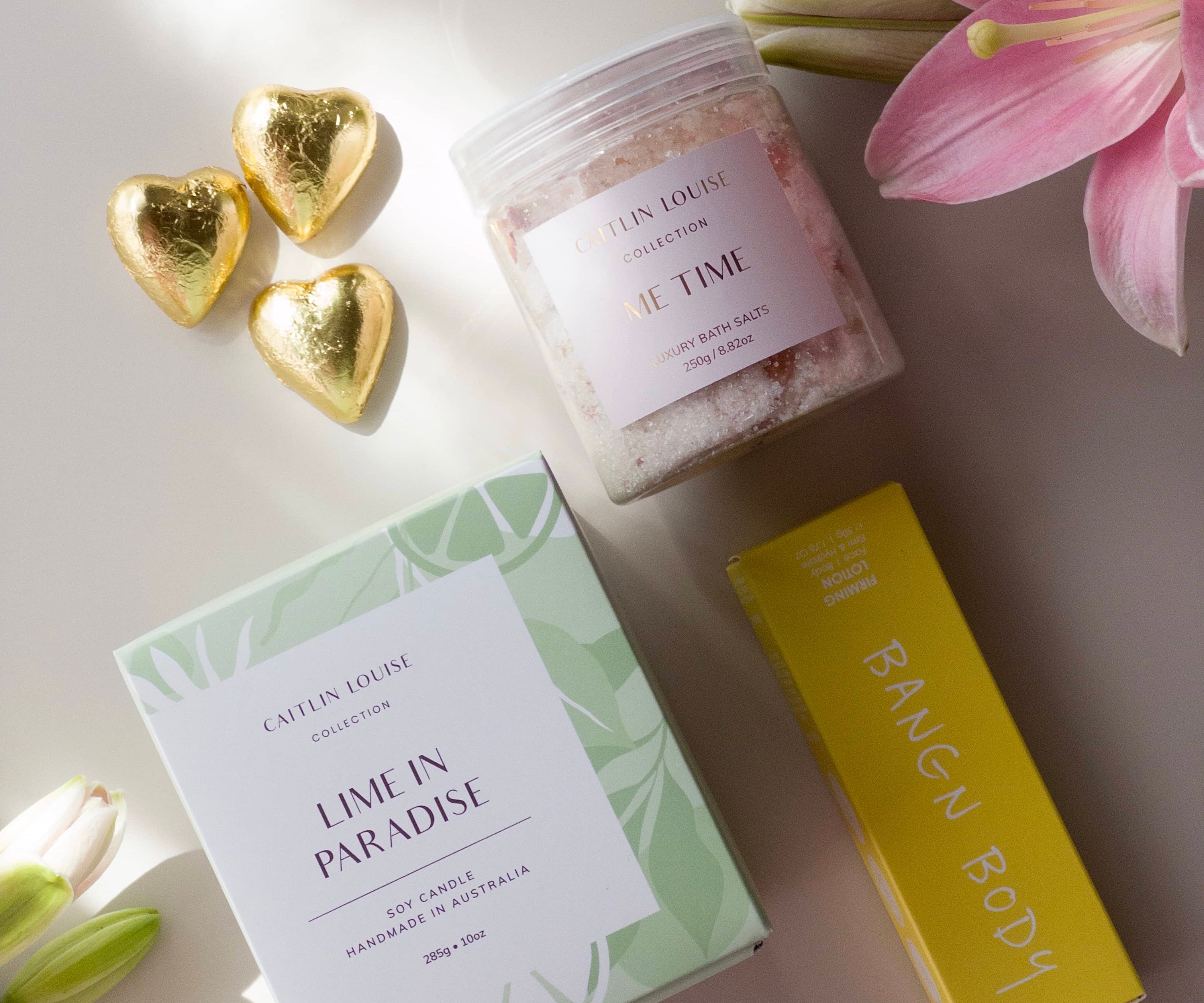 Flat lay of gift bundle for pampering - lime in paradise candle, me time bath salts, bangn body firming lotion and three heart shaped chocolates - surrounded by flowers.