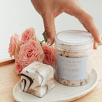 Hand reaching for Caitlin Louise Collection Unwind Luxury Bath Salts. Jar sitting on tray with flowers behind it.