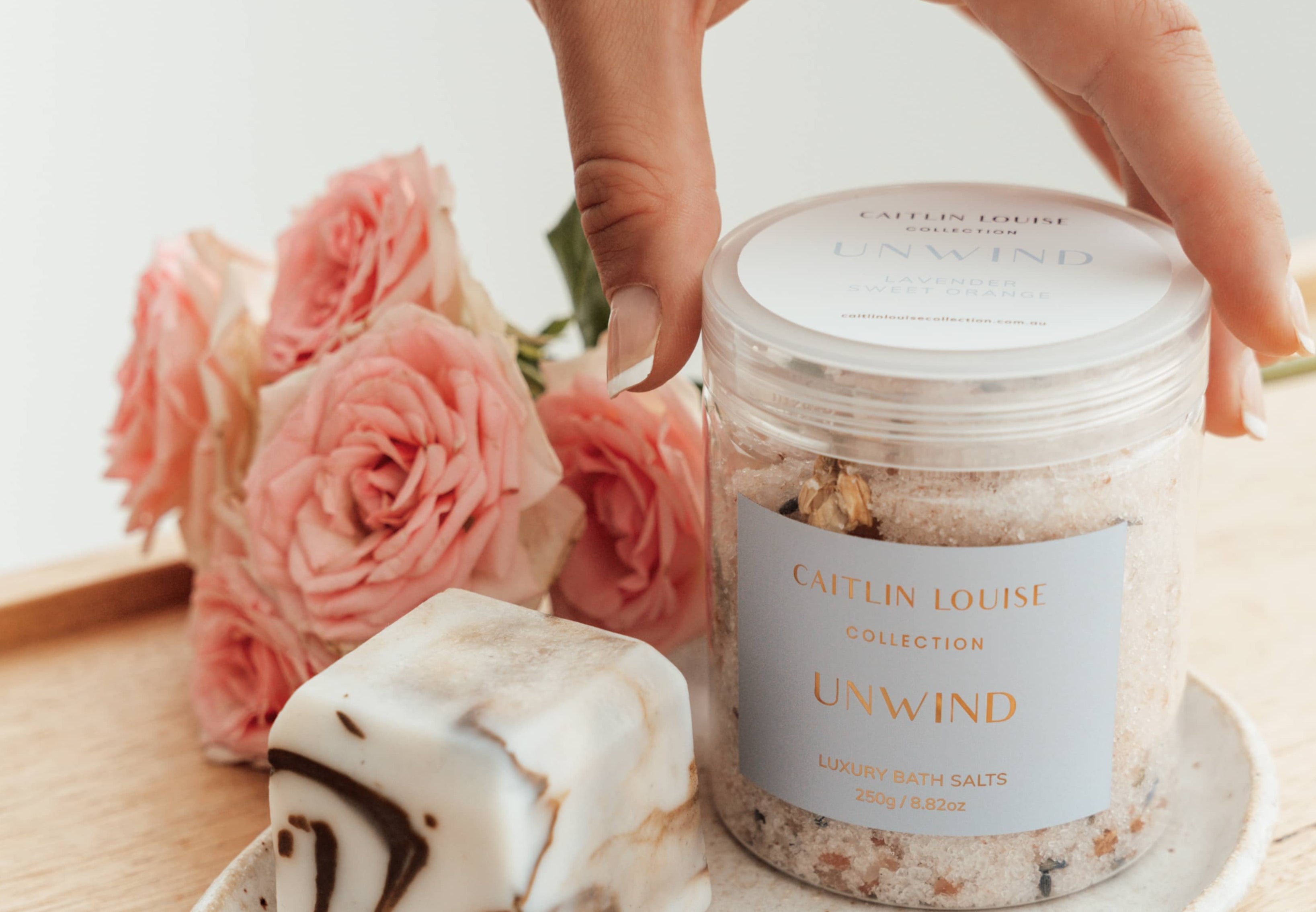 Hand reaching for Caitlin Louise Collection Unwind Luxury Bath Salts. Jar sitting on tray with flowers behind it.