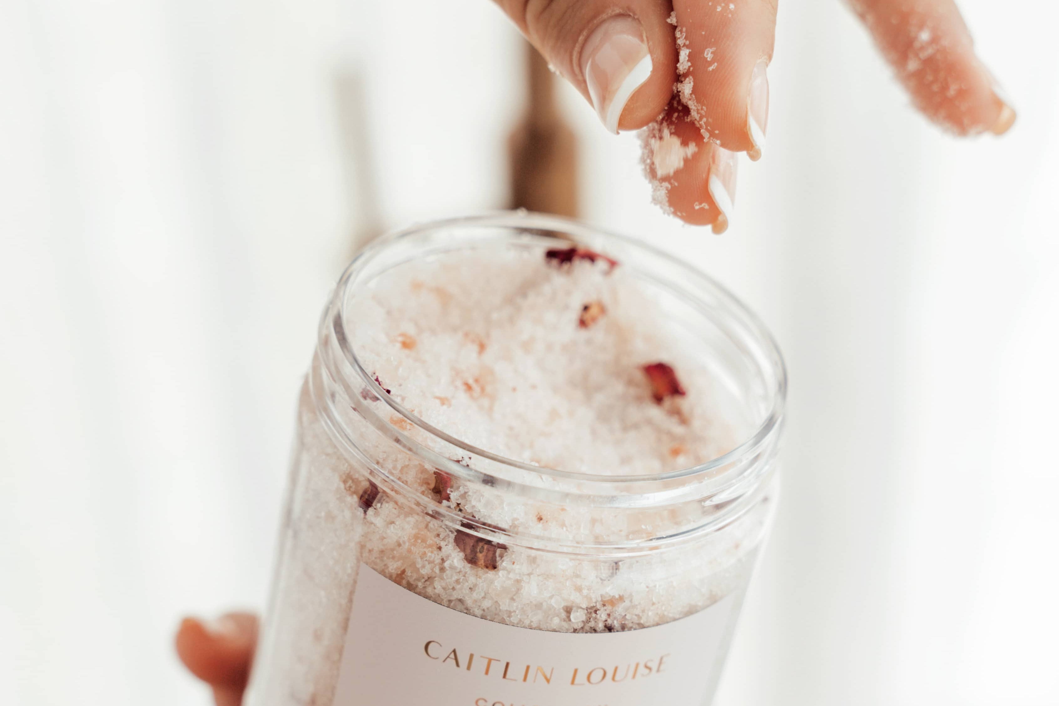 Caitlin Louise Collection Me Time luxury bath salts, with rose petals in the jar. Hand scooping some salts out of the jar, front view.