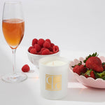 Glass of pink champagne, bowl of raspberries, and bowl of strawberries with a Caitlin Louise Collection candle on a white background