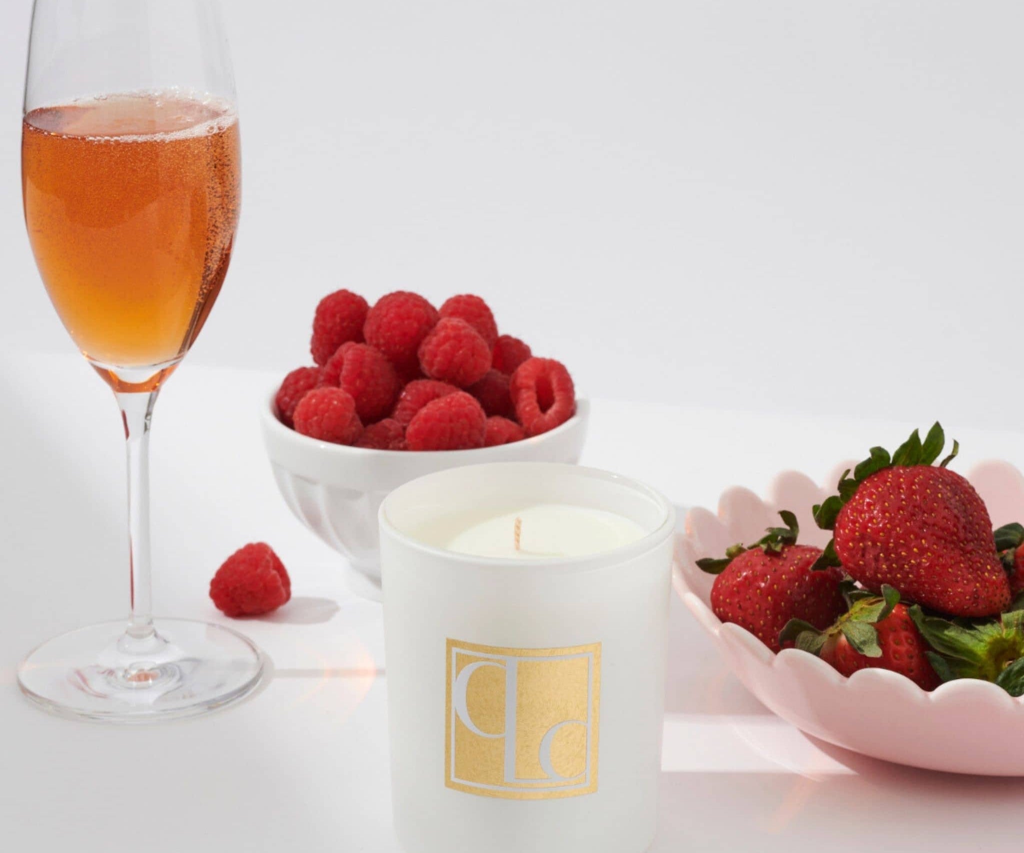 Glass of pink champagne, bowl of raspberries, and bowl of strawberries with a Caitlin Louise Collection candle on a white background