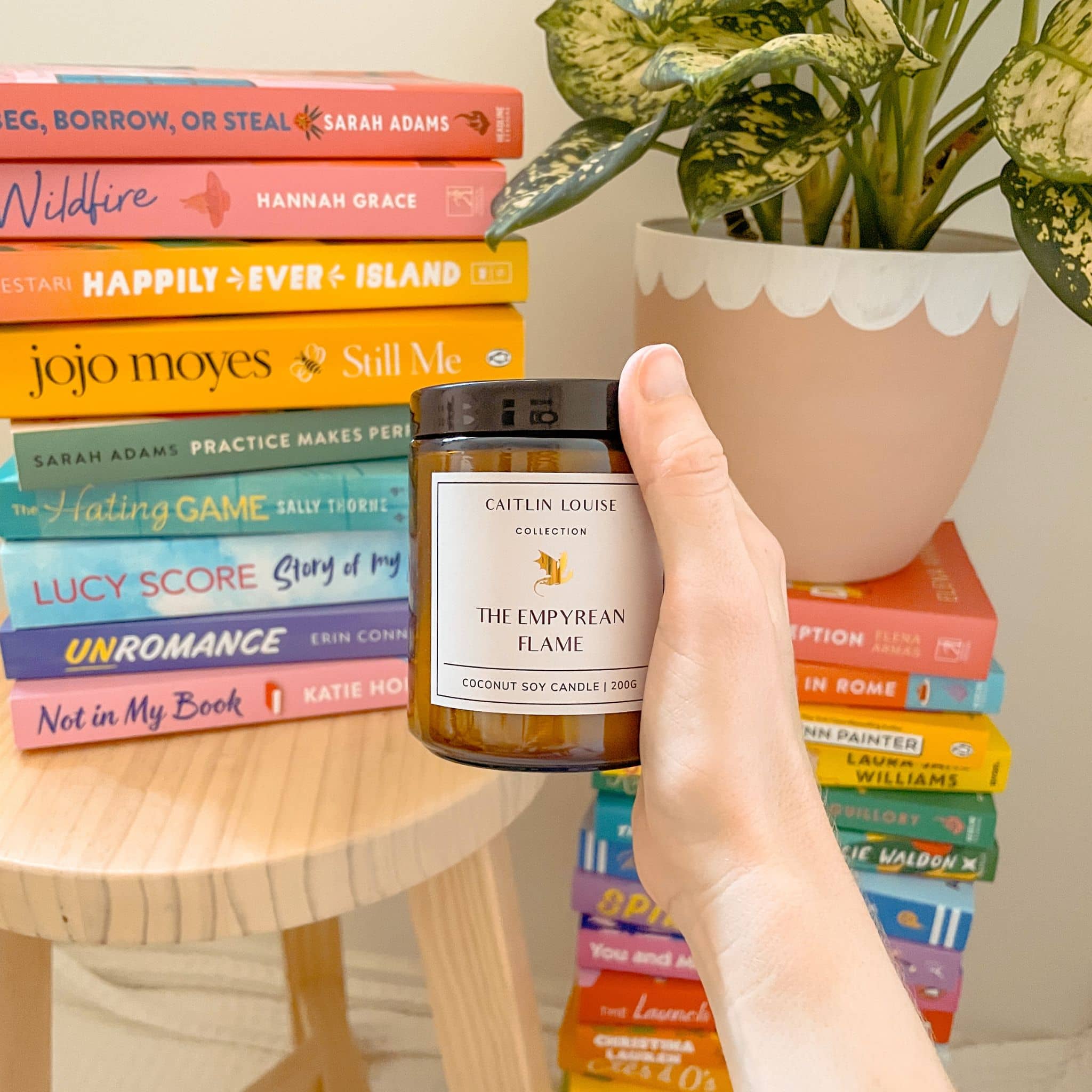 Hand holding a candle labeled 'The Empyrean Flame' in front of a stack of books and a plant.