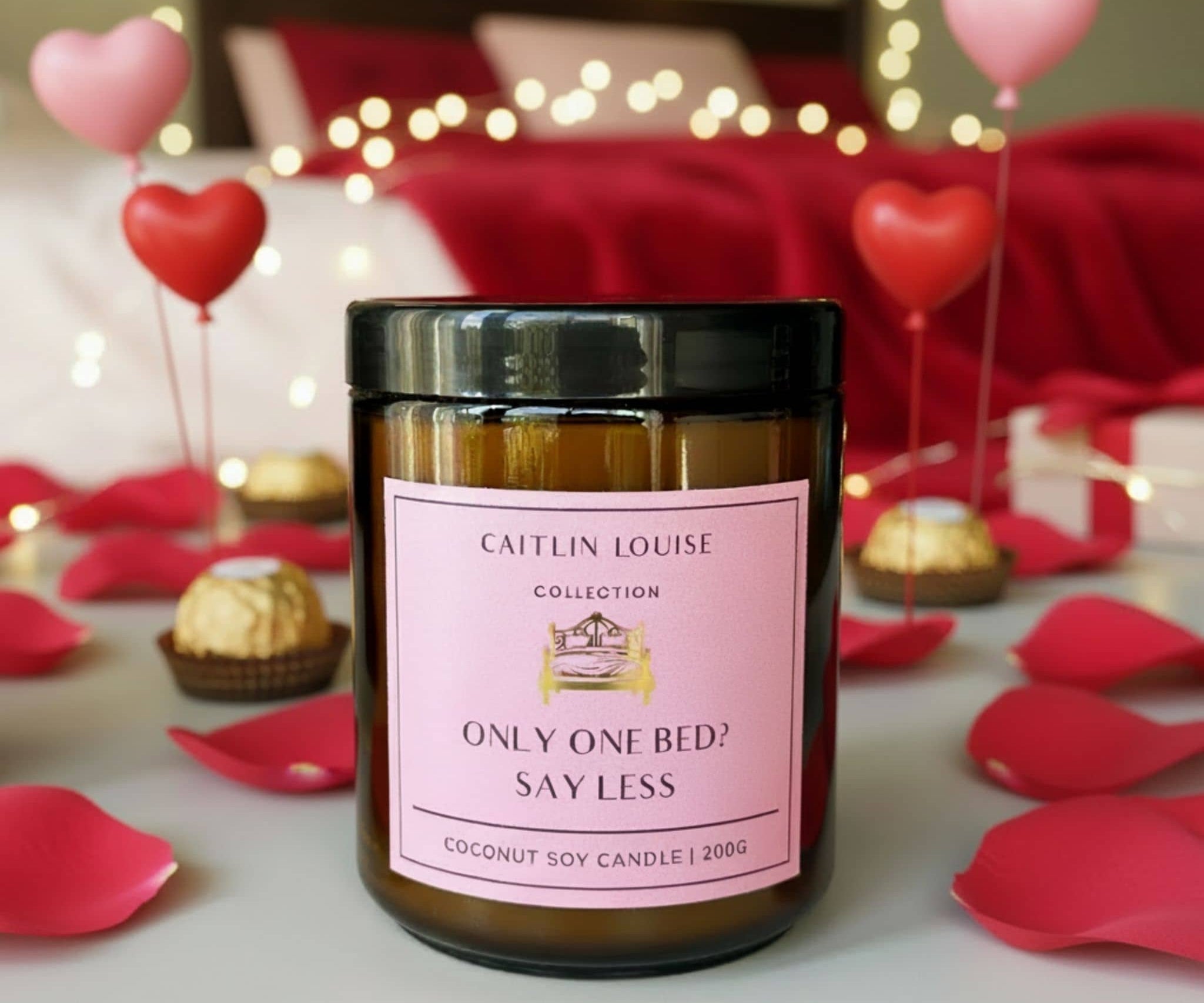 Candle with a pink label in a romantic setting with heart balloons and rose petals.