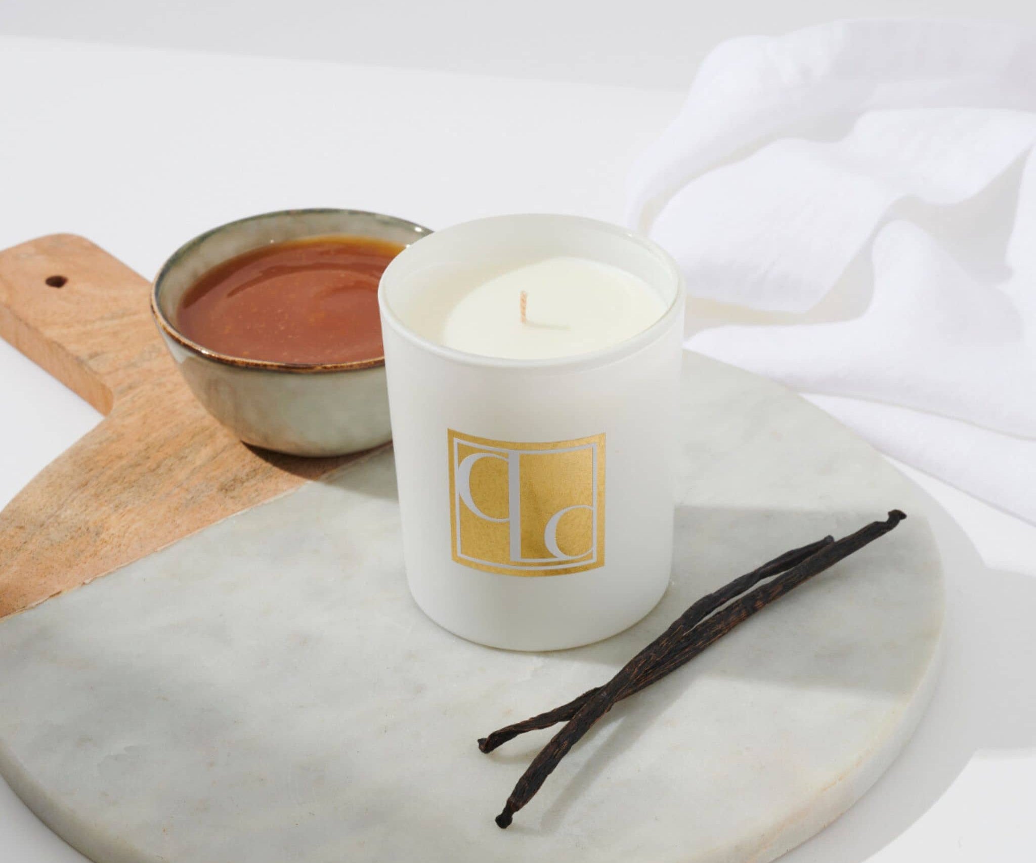 Caitlin Louise Collection Vanilla Caramel Scented Candle on board with bowl of caramel in background and vanilla sticks in the foreground.