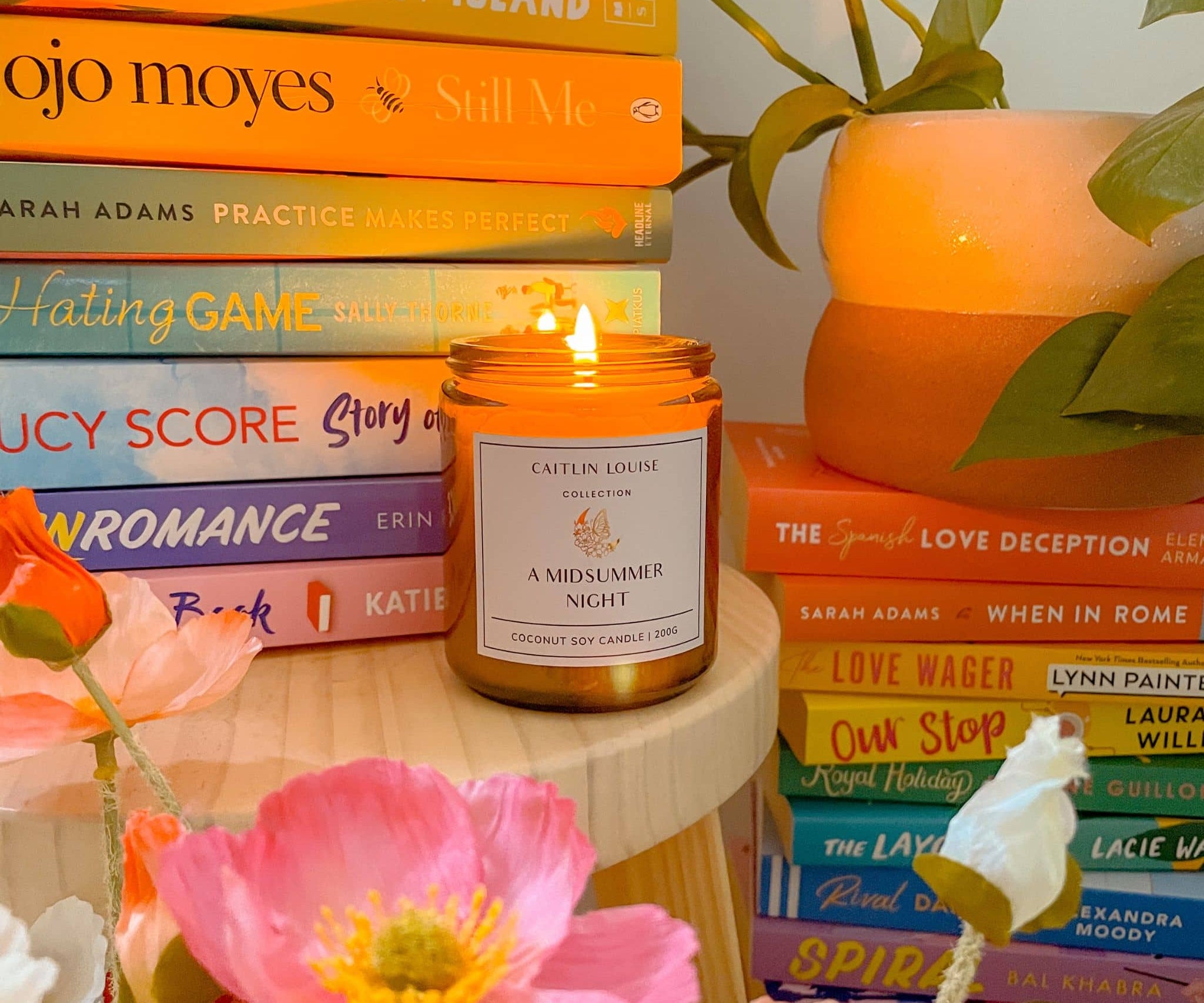 Candle titled 'A Midsummer Night' on a stack of books with flowers and a plant in the background.