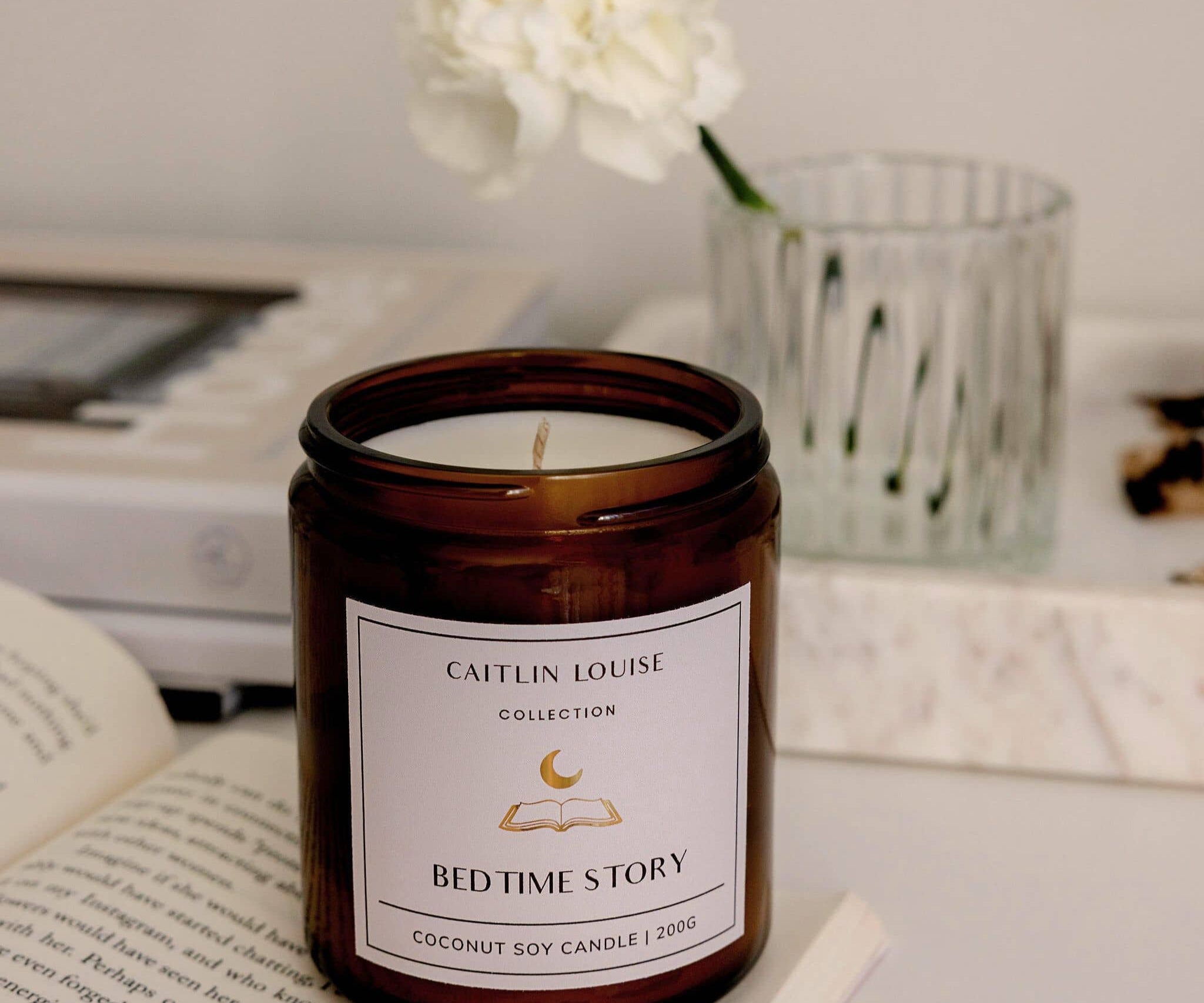 Candle labeled 'Caitlin Louise Collection Bed Time Story' on a surface with books and flowers in the background.