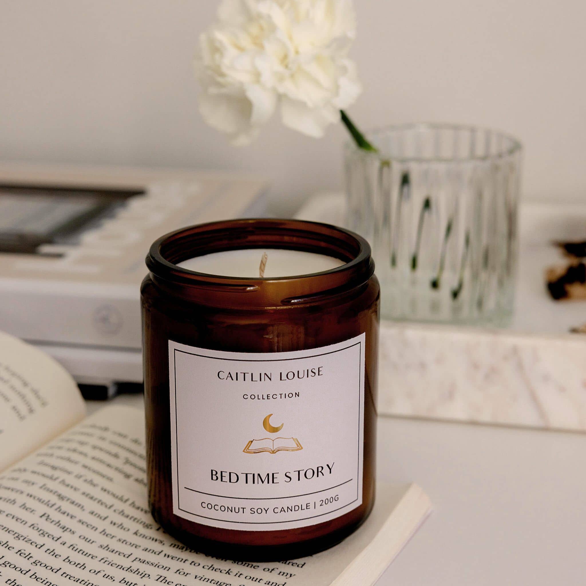 Candle labeled 'Caitlin Louise Collection Bed Time Story' on a surface with books and flowers in the background.