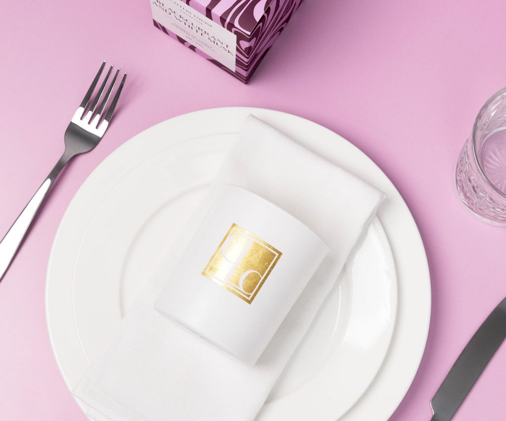 Caitlin Louise Collection candle with gold logo on a white plate with cutlery on a pink background