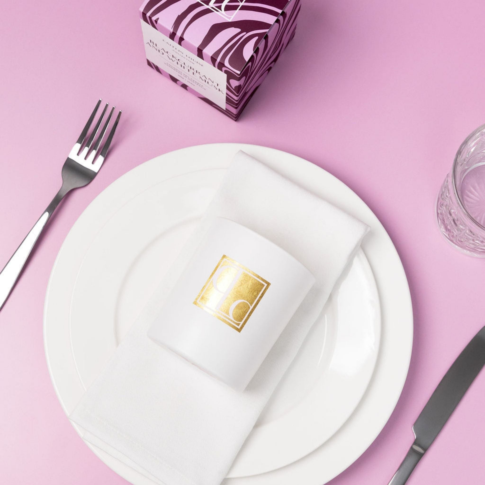 Caitlin Louise Collection candle with gold logo on a white plate with cutlery on a pink background