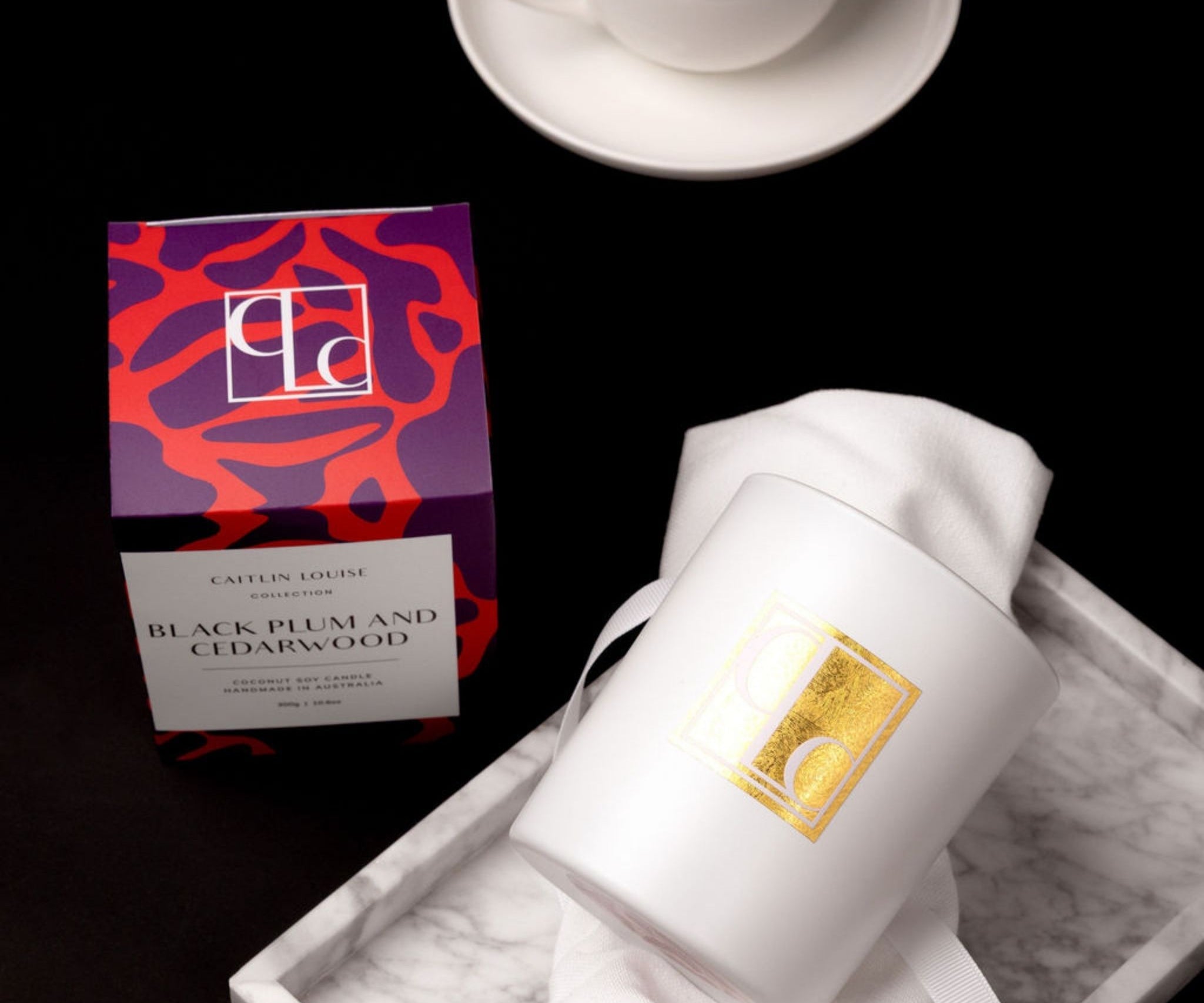 Caitlin Louise collection candle with colourful packaging on a marble tray, cup of tea in background on black background