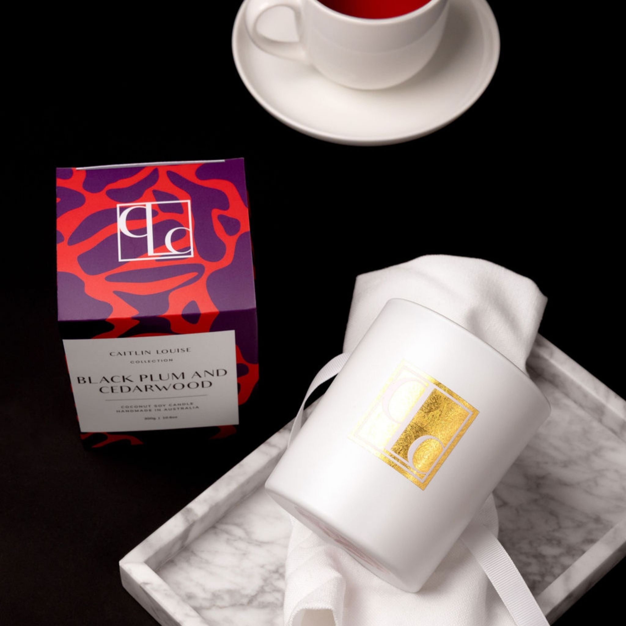Caitlin Louise collection candle with colourful packaging on a marble tray, cup of tea in background on black background