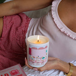 Person holding a lit candle with a decorative label, wearing a pink shirt and red checkered shorts.