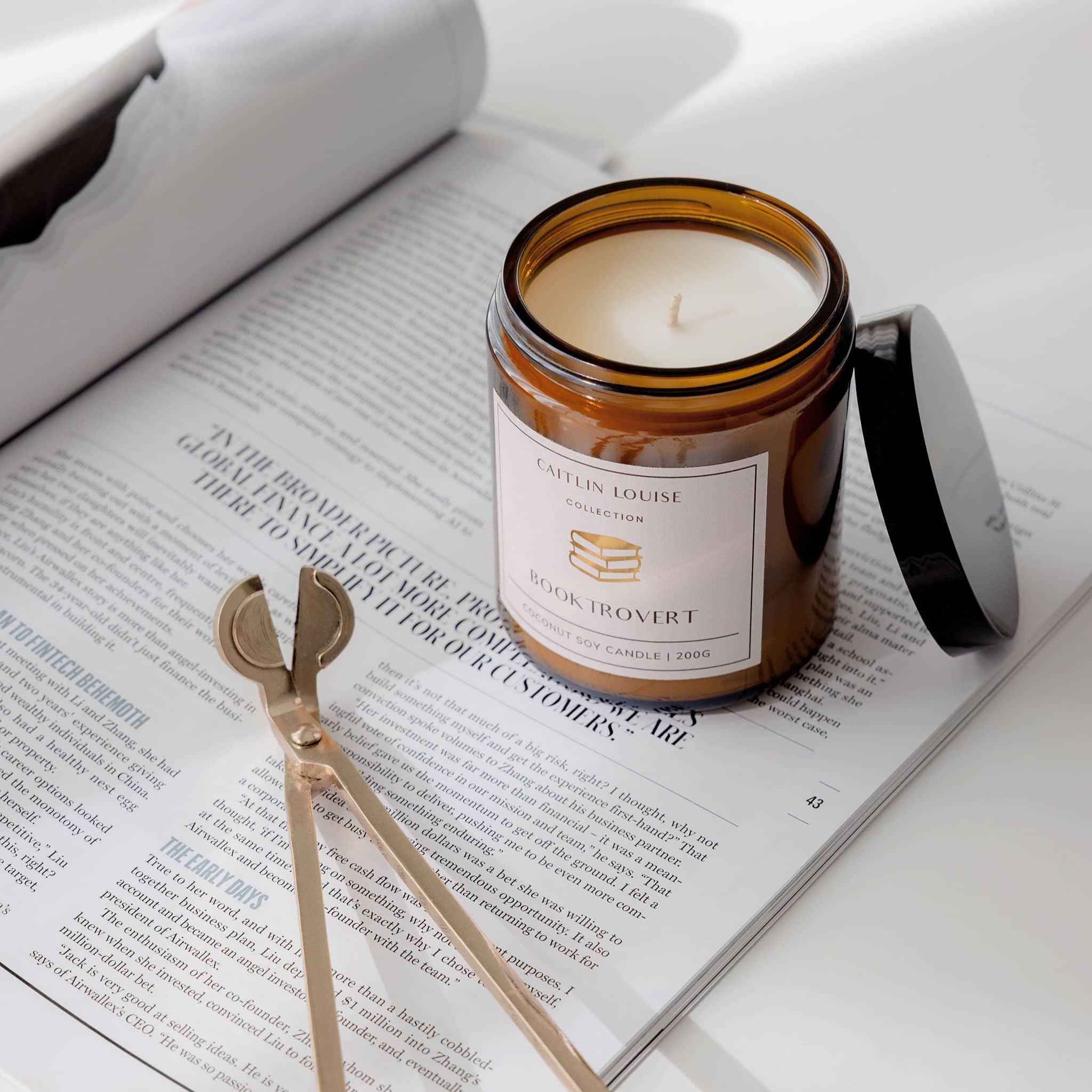 Candle in a glass jar labeled 'Booktrovert' on an open book with a pair of wick trimmers.