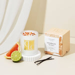 Candle labeled 'Champagne Sunset' with packaging, fruit, and vanilla beans on a neutral background