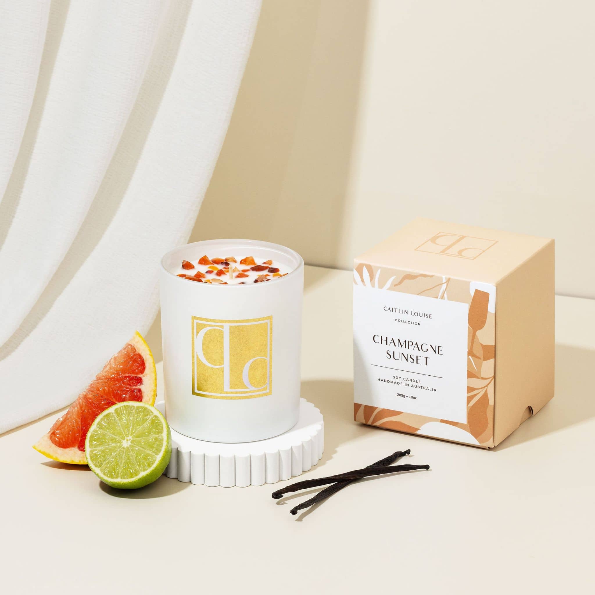 Candle labeled 'Champagne Sunset' with packaging, fruit, and vanilla beans on a neutral background