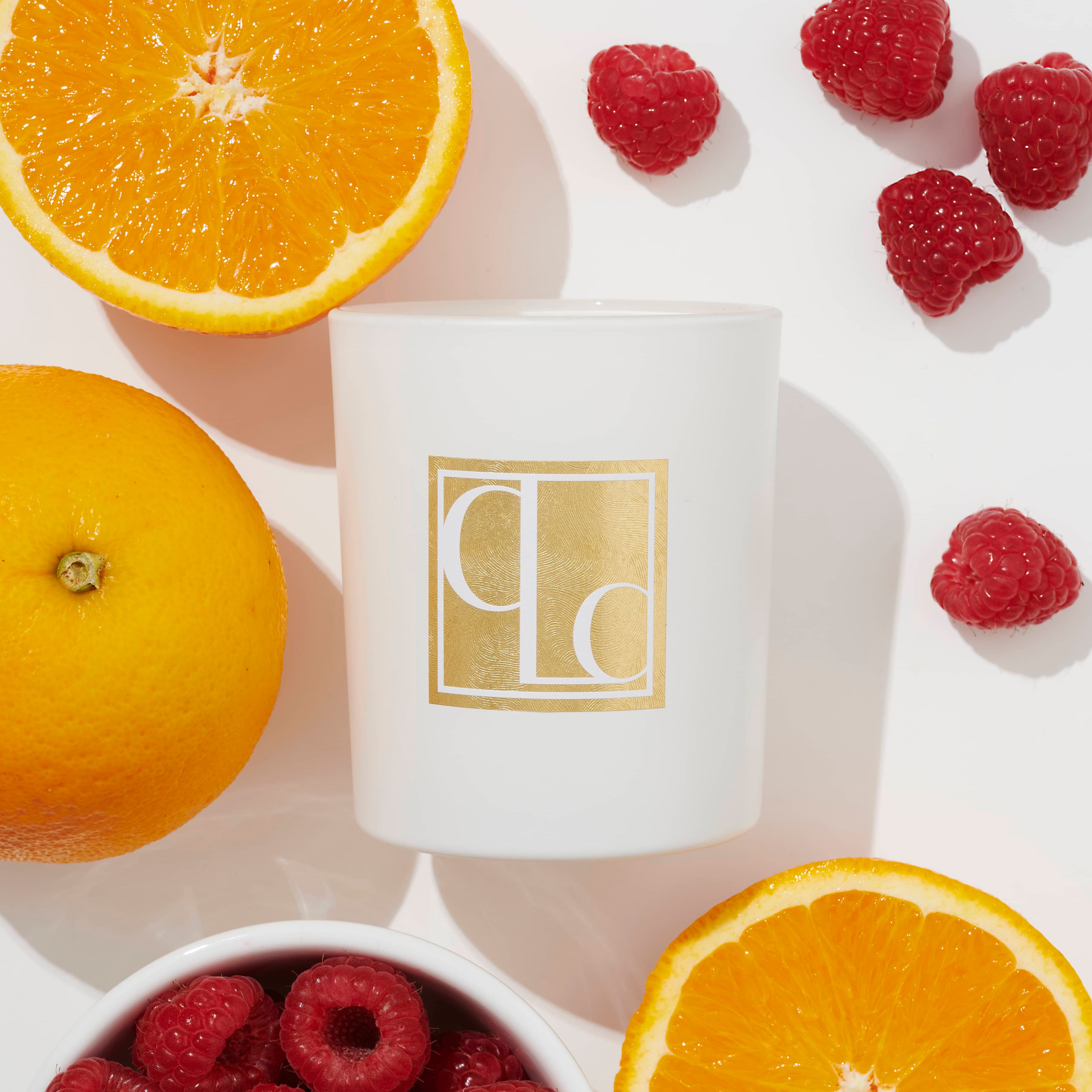 A white Caitlin Louise Collection candle with a gold label, surrounded by orange slices and raspberries, on a white background.