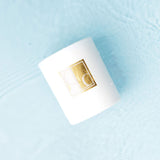 White candle with a gold logo on a light blue surface covered in water