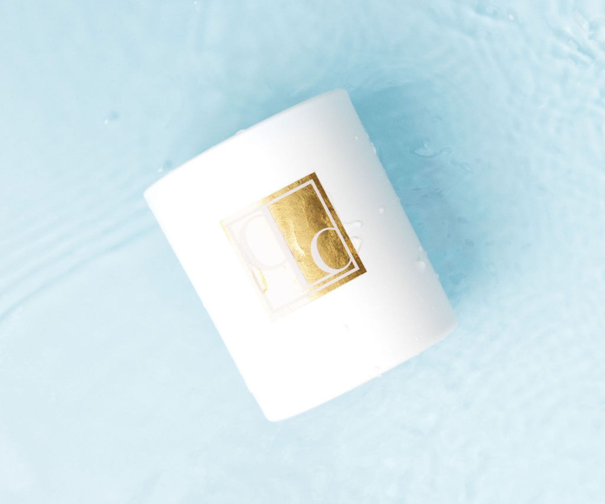 White candle with a gold logo on a light blue surface covered in water
