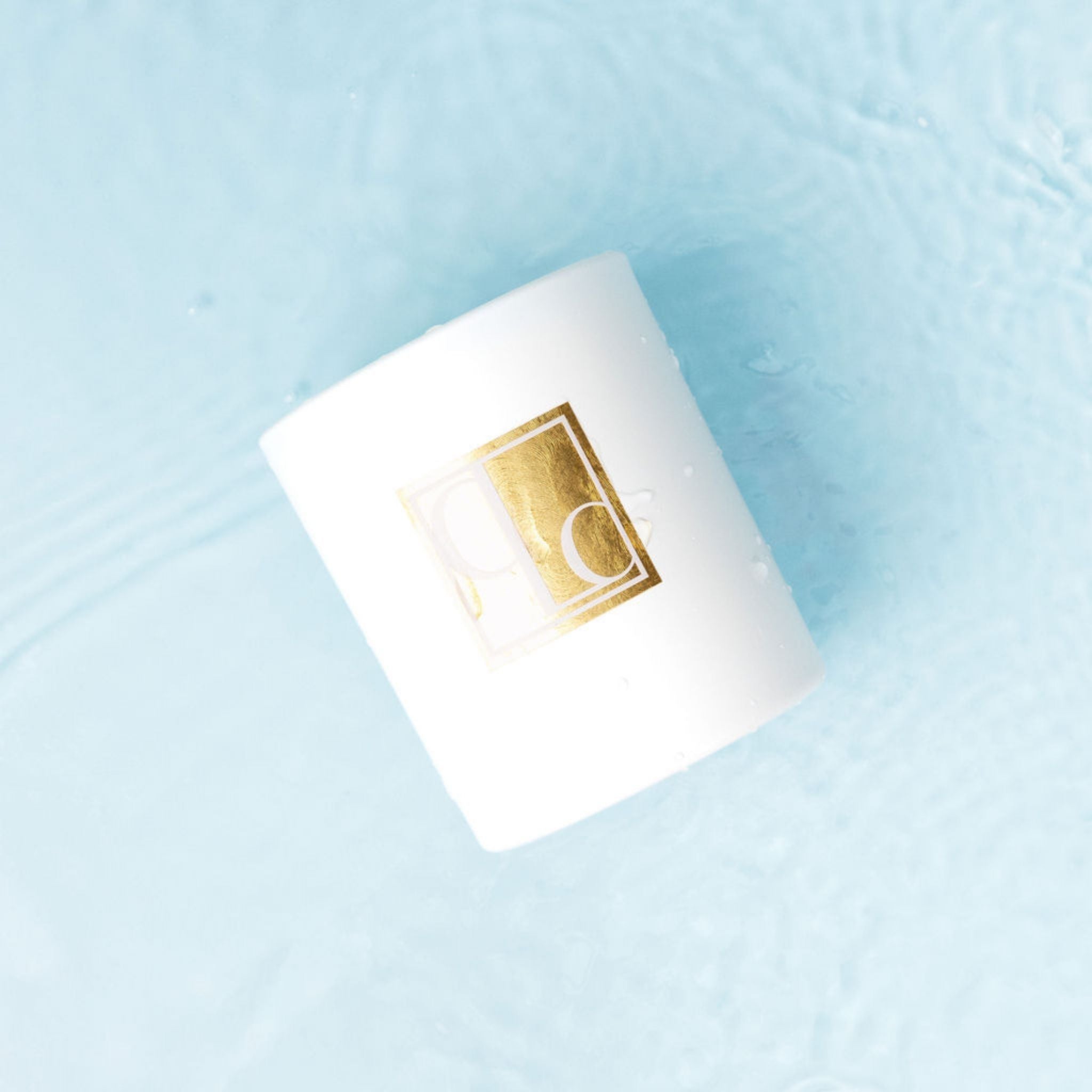 White candle with a gold logo on a light blue surface covered in water