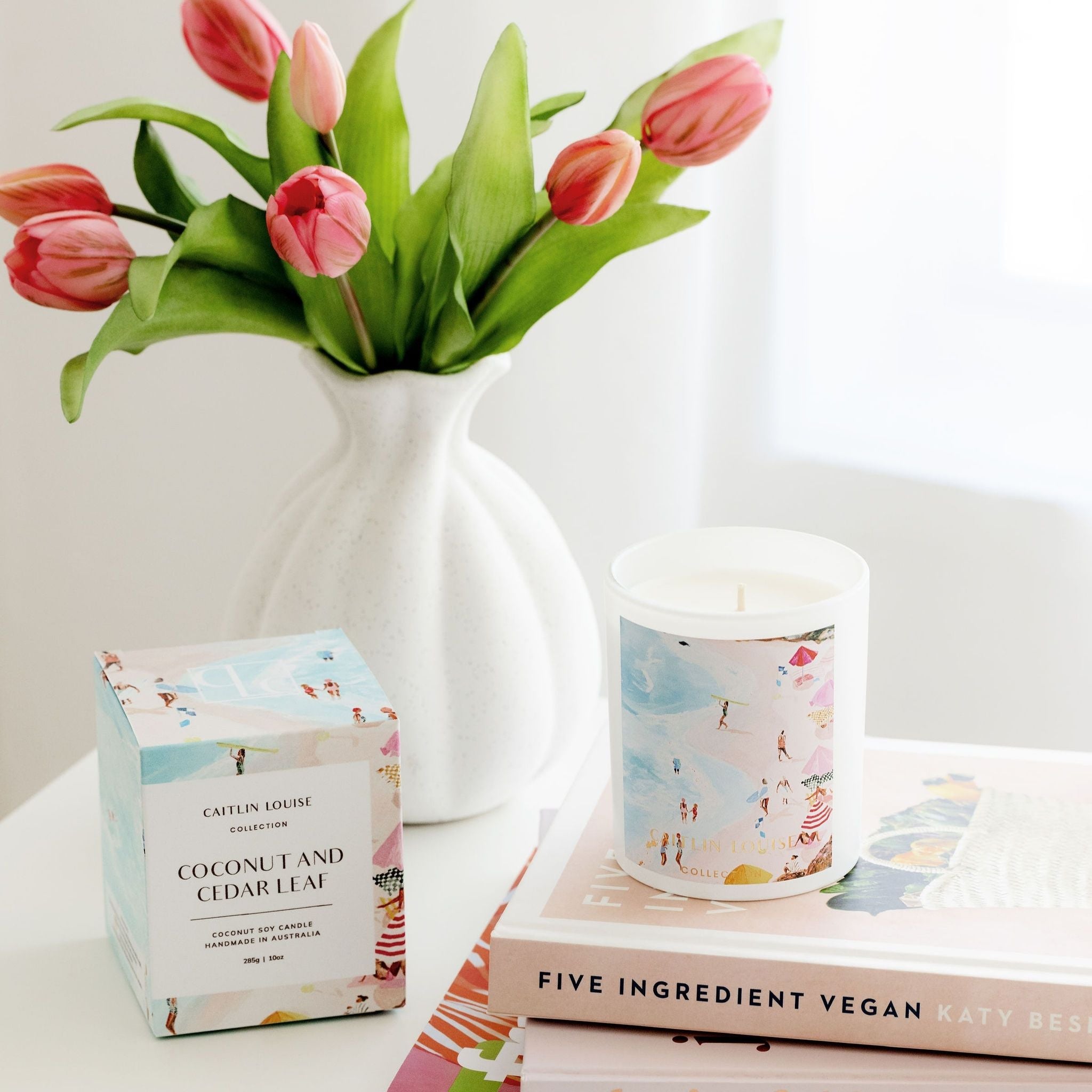 Candle and packaging with a vase of pink tulips on a white surface