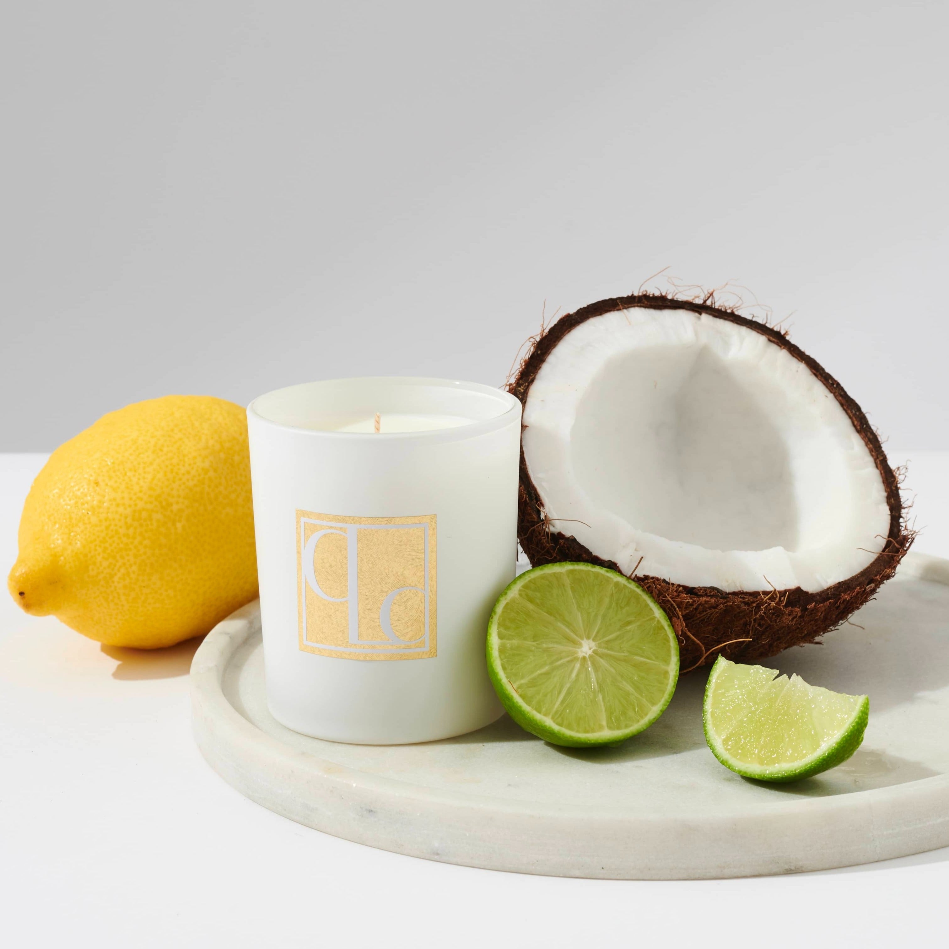 A white candle with the scent of coconut lime, placed on a marble surface, accompanied by a halved coconut, a lemon, and lime slices.