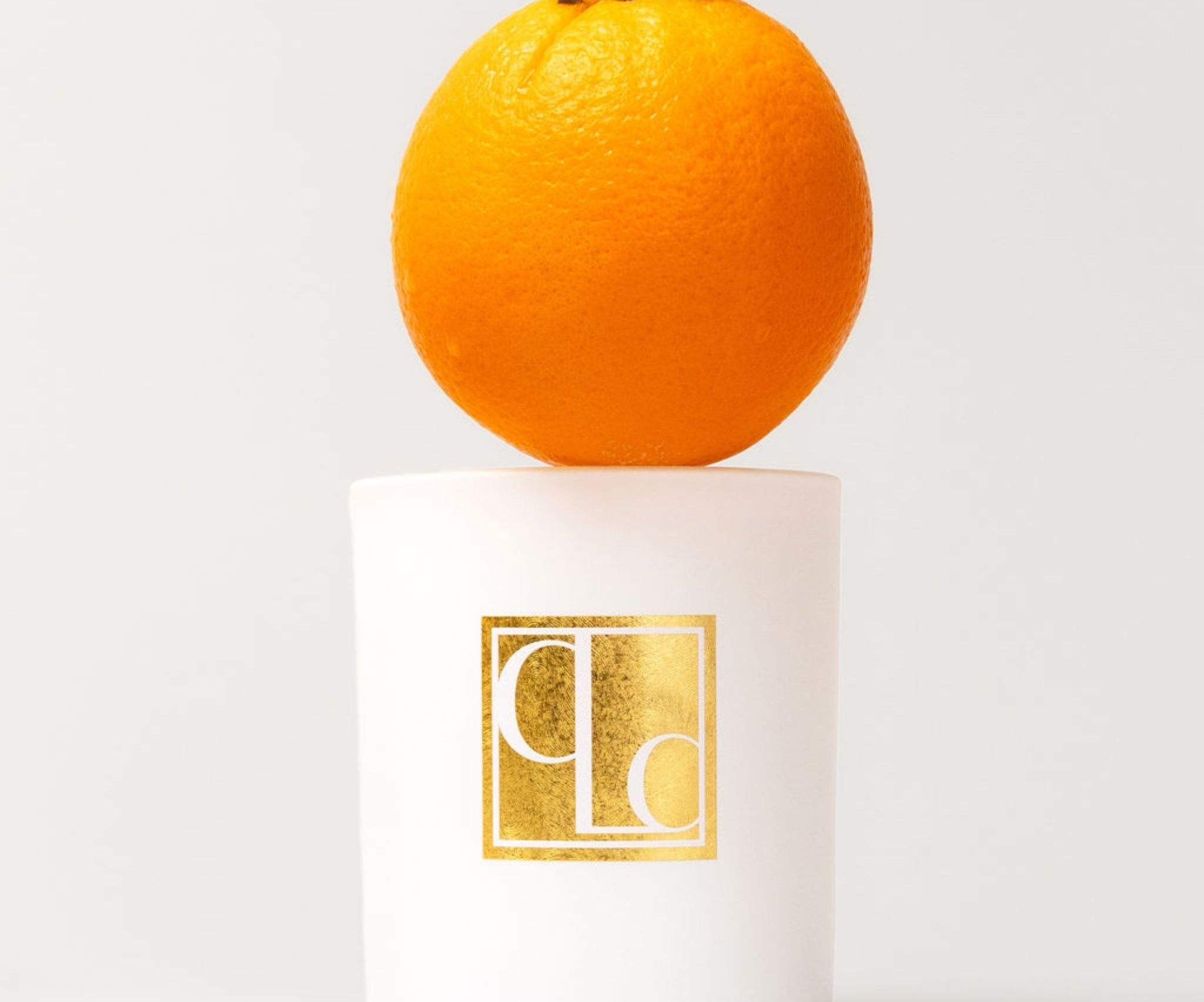 Candle with a gold logo on a white base, featuring an orange on top against a light gray background.