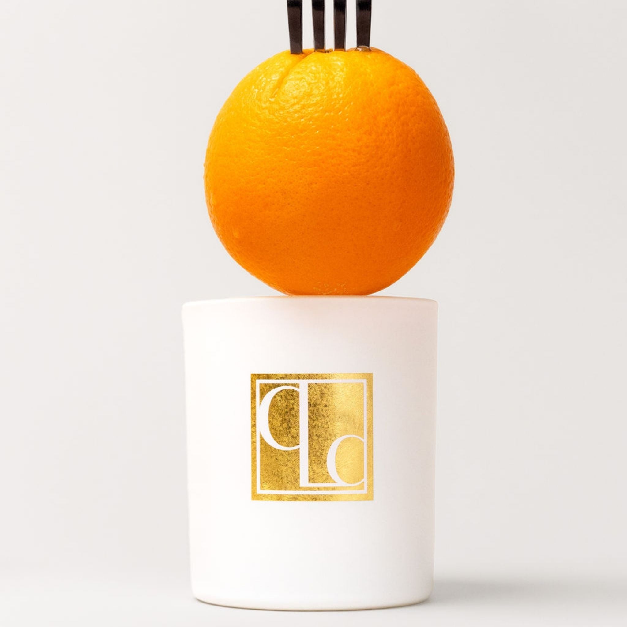 Candle with a gold logo on a white base, featuring an orange on top against a light gray background.