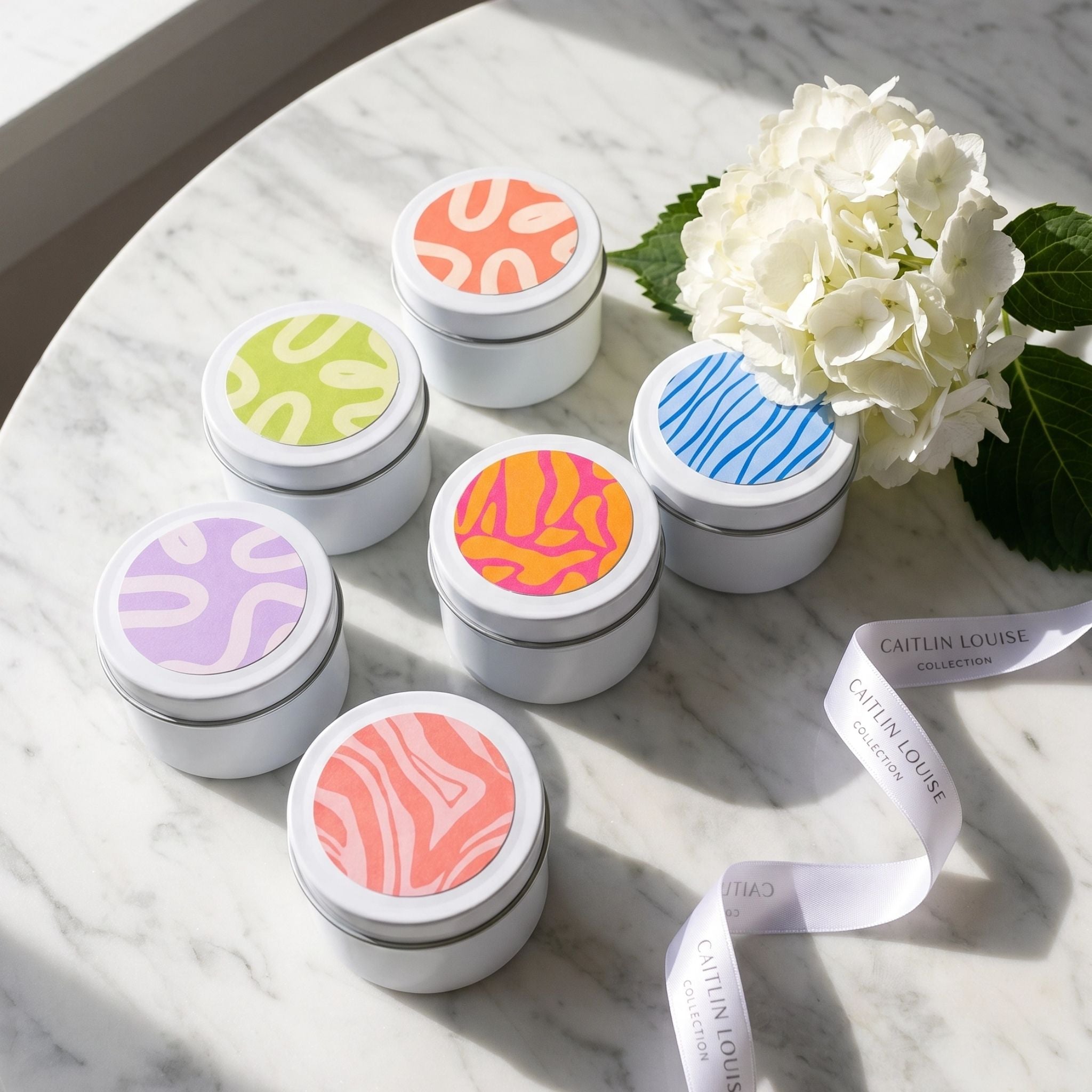 Set of small discovery set candle tins with colourful lids on a marble surface with a white flower.