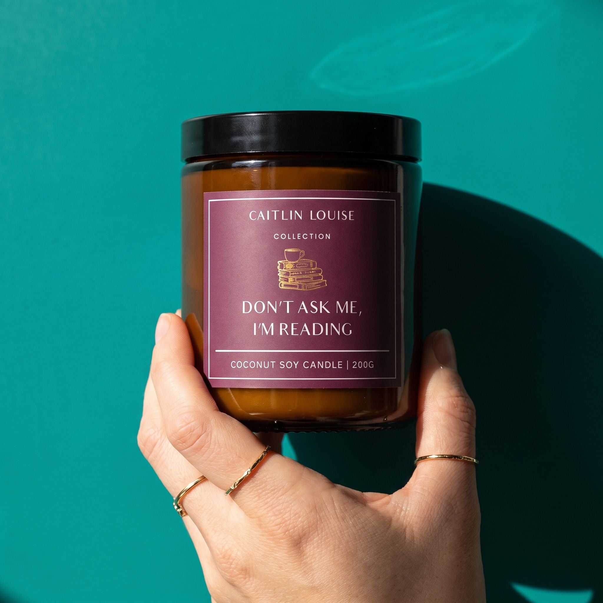 hand holding candle with purple label saying 'don't ask me, i'm reading' against aqua coloured background