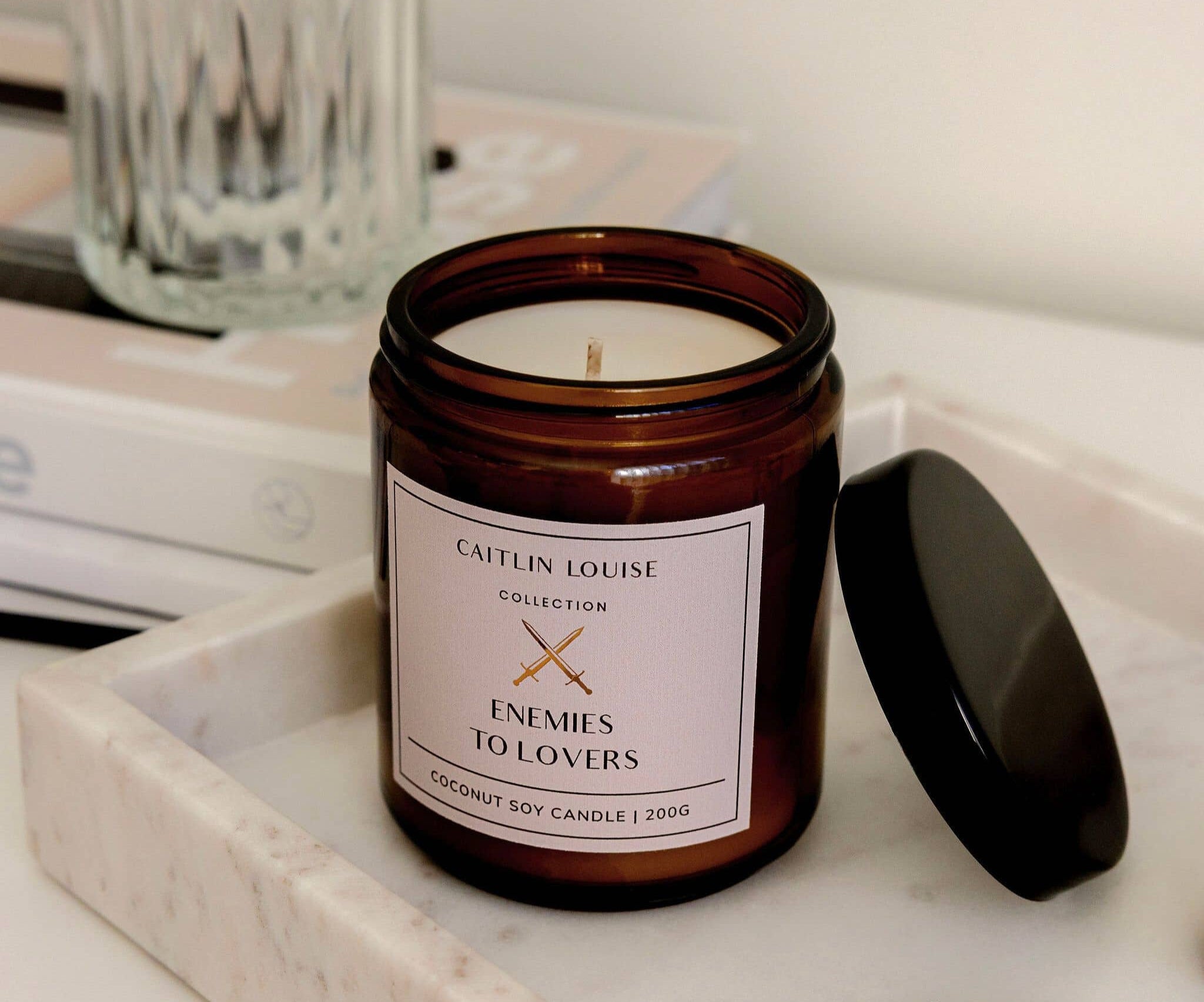 Candle jar labeled 'Enemies to Lovers' by Caitlin Louise Collection on a marble surface.