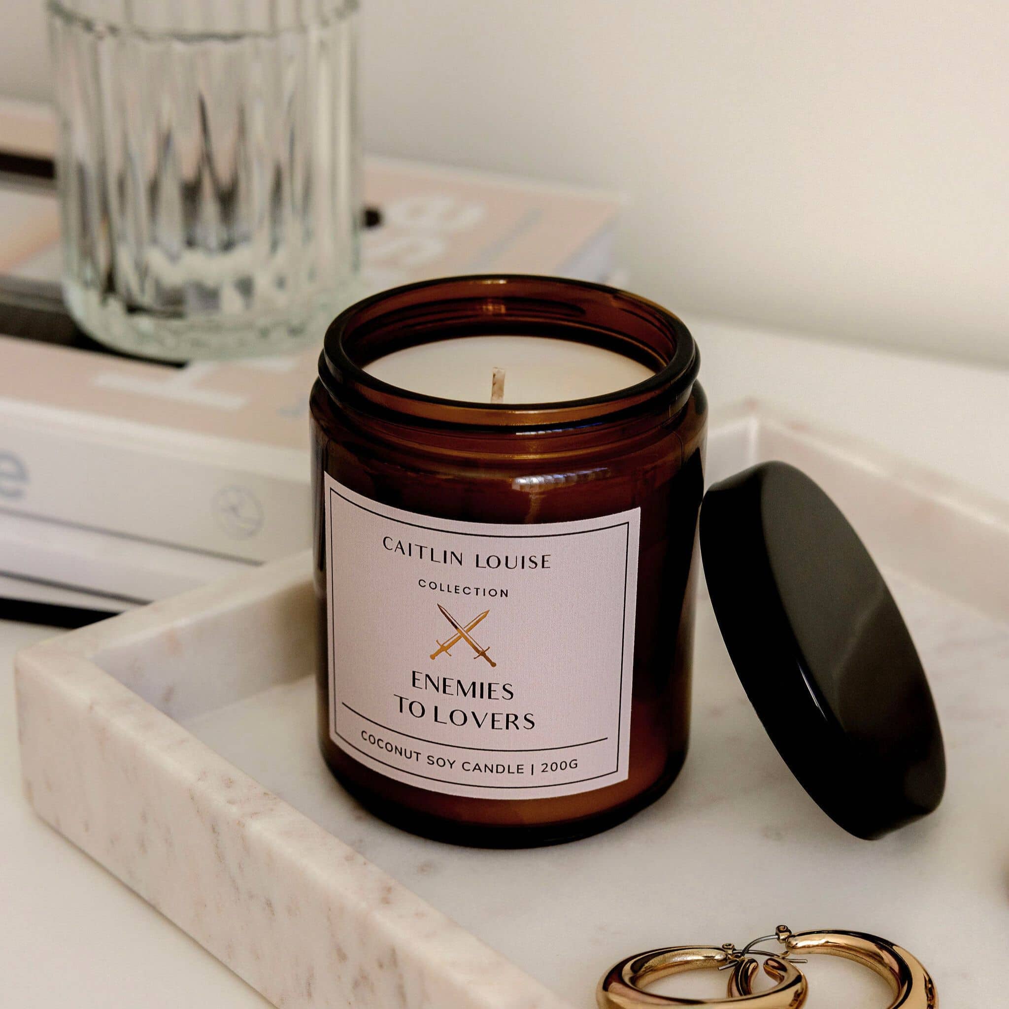 Candle jar labeled 'Enemies to Lovers' by Caitlin Louise Collection on a marble surface.