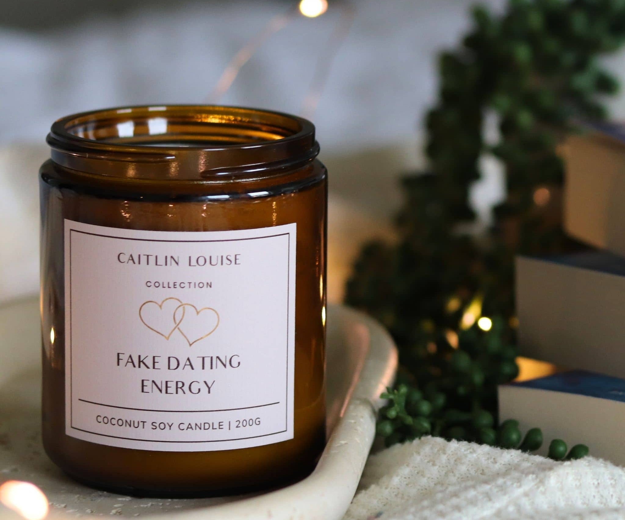 Candle with a label 'Fake Dating Energy' by Caitlin Louise Collection, surrounded by decorative elements.
