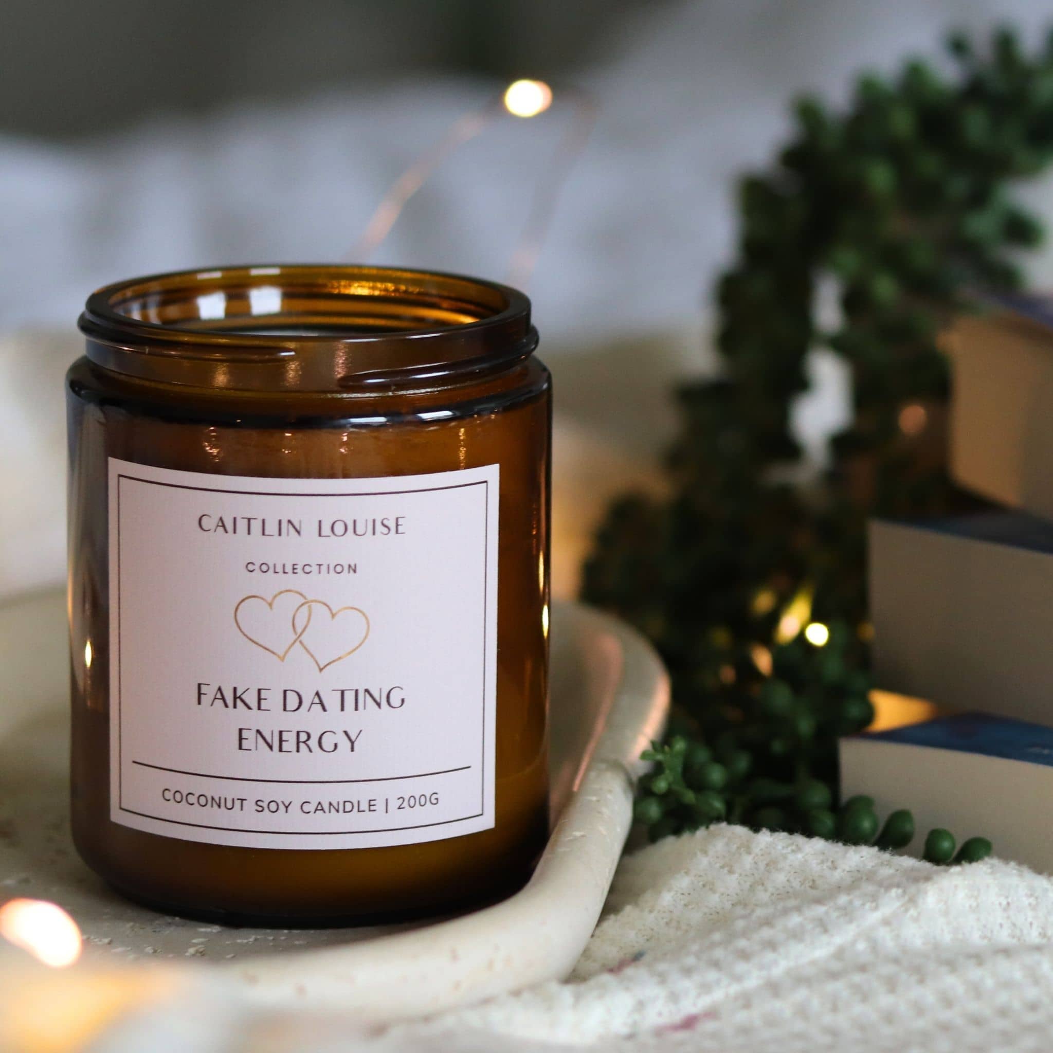 Candle with a label 'Fake Dating Energy' by Caitlin Louise Collection, surrounded by decorative elements.