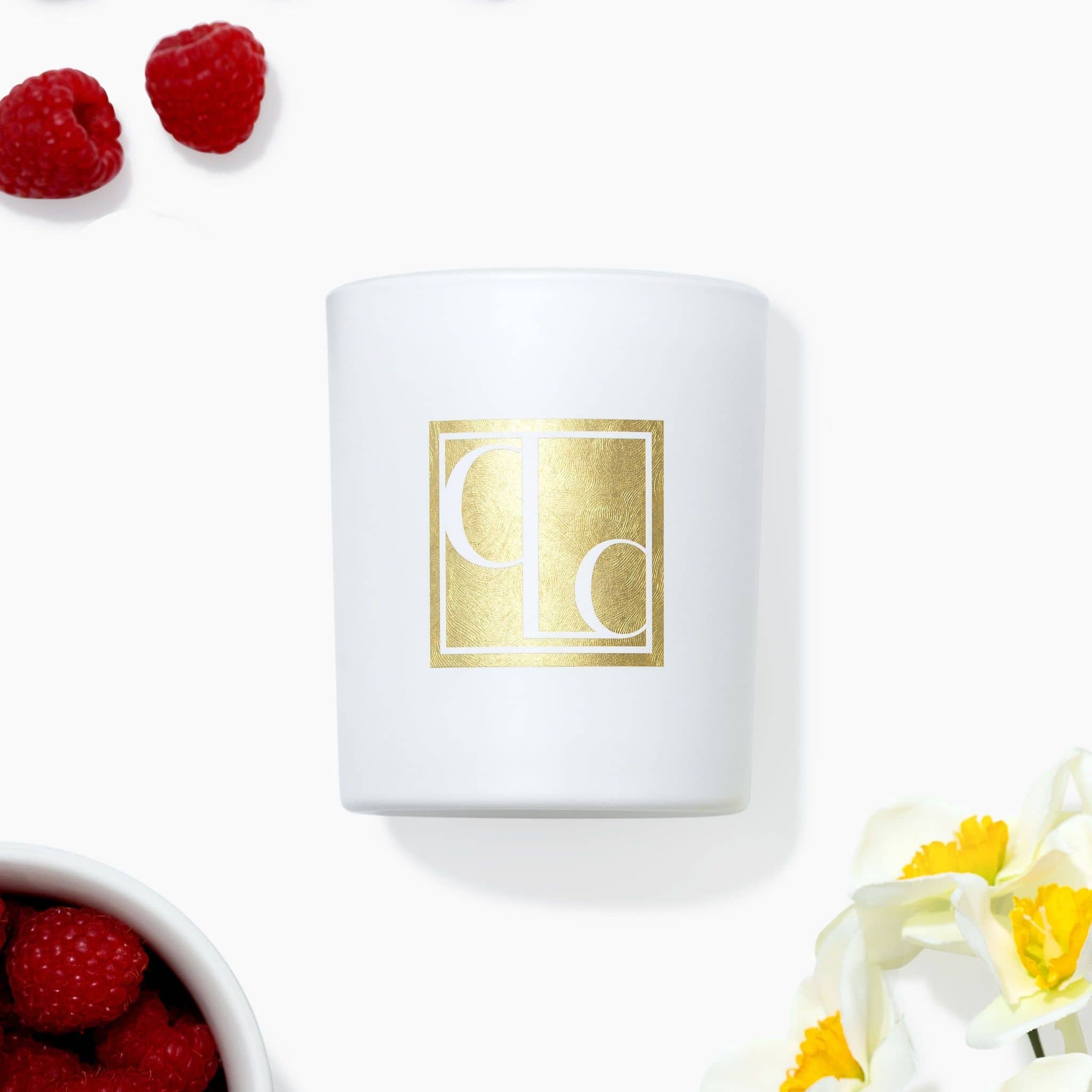 White candle with gold logo surrounded by raspberries and flowers on a white background
