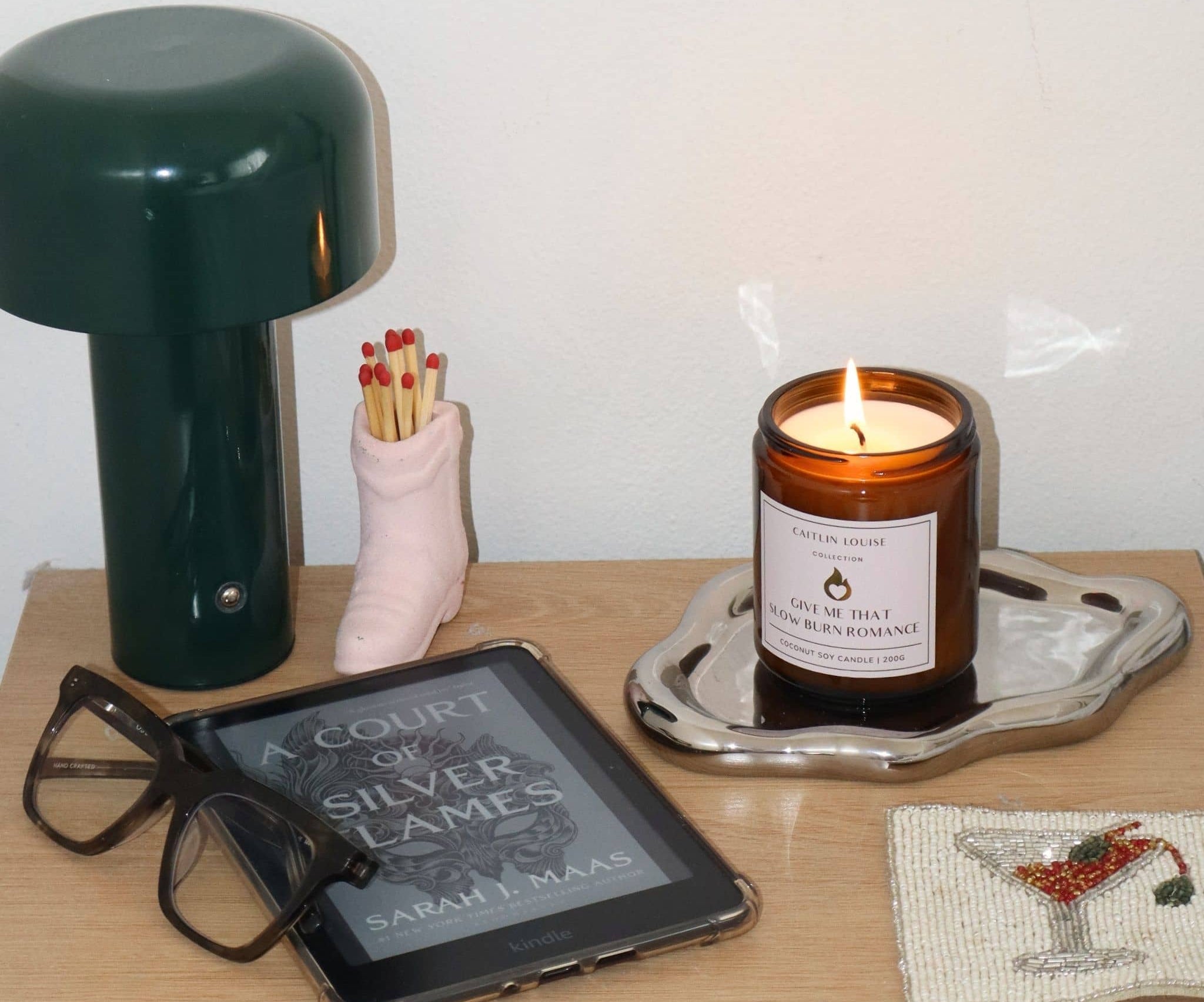 Tabletop scene with a lit candle, book, glasses, and small lamp.